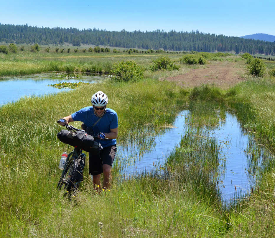 In this June 7, 2013 photo, Donnie Kolb pushes his bike through a marshy section of Sycan Marsh near Klamath Falls, Ore.  Kolb has been organizing gravel bicycle rides for the past five years, usually keeping his events a bit on the down low.  His outings are unsupported, unsanctioned, have no entry fee and crown no winner. (AP Photo/The Bulletin, Gabriel Amadeus)