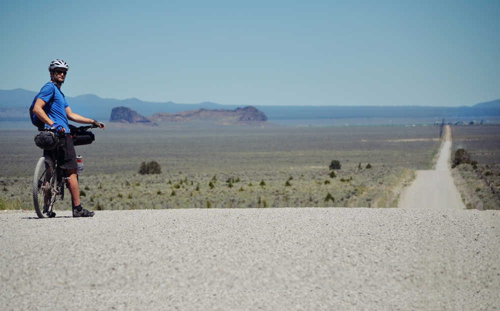 In this June 8, 2013 photo, Donnie Kolb pauses on a long stretch of gravel road with central Oregon's Fork Rock formation visible in the distance. Kolb has been organizing gravel bicycle rides for the past five years, usually keeping his events a bit on the down low.  His outings are unsupported, unsanctioned, have no entry fee and crown no winner. (AP Photo/The Bulletin, Gabriel Amadeus)