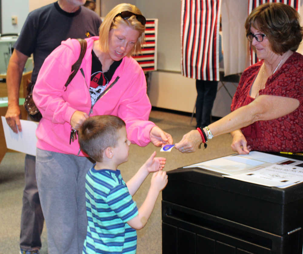 Photo by Dan Balmer/Peninsula Clarion Diane Hinshaw hands a sticker to five-year-old Urijah Walden and his mother Tasha Walden after voting in the primary election at Soldotna City Hall Tuesday.