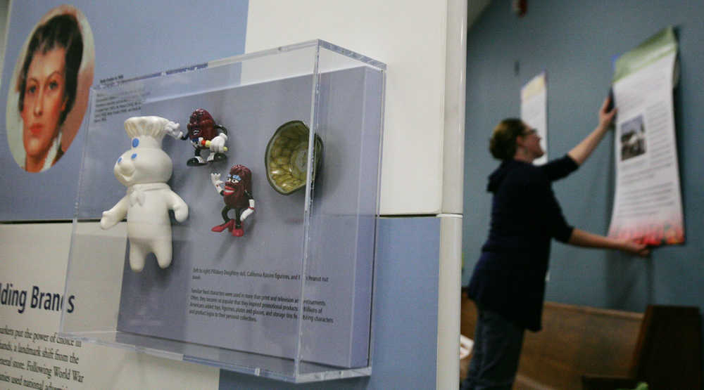 In this photo taken on Thursday, Oct. 2, 2014, Tamara Martz, exhibit and graphic designer at the University of Alaska Museum of the North, hangs wall boards in the background as a case including the Pillsbury Doughboy doll, California Raisins figurines and Mr. Peanut nut bowl is on display as the "Key Ingredients: America By Food" Smithsonian Institution traveling exhibit is installed at the UAF Community and Technical College on Barnette Street in Fairbanks, Alaska. (AP Photo/Fairbanks Daily News-Miner, Eric Engman)