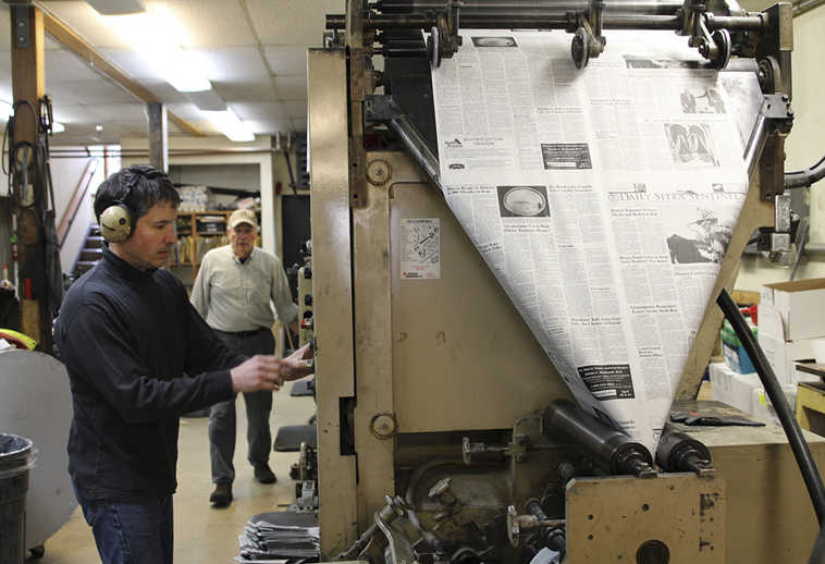 In this April 15, 2015 photo, James Poulson, left, operates the printing press as his father, Thad, approaches at the Daily Sitka Sentinel newspaper office in Sitka, Alaska. As many newspapers in big cities have folded or turned into online only operations, the Daily Sitka Sentinel, which celebrated its 75th anniversary last year, steadily churns out five issues a week. (Emily Kwong/KCAW via AP)
