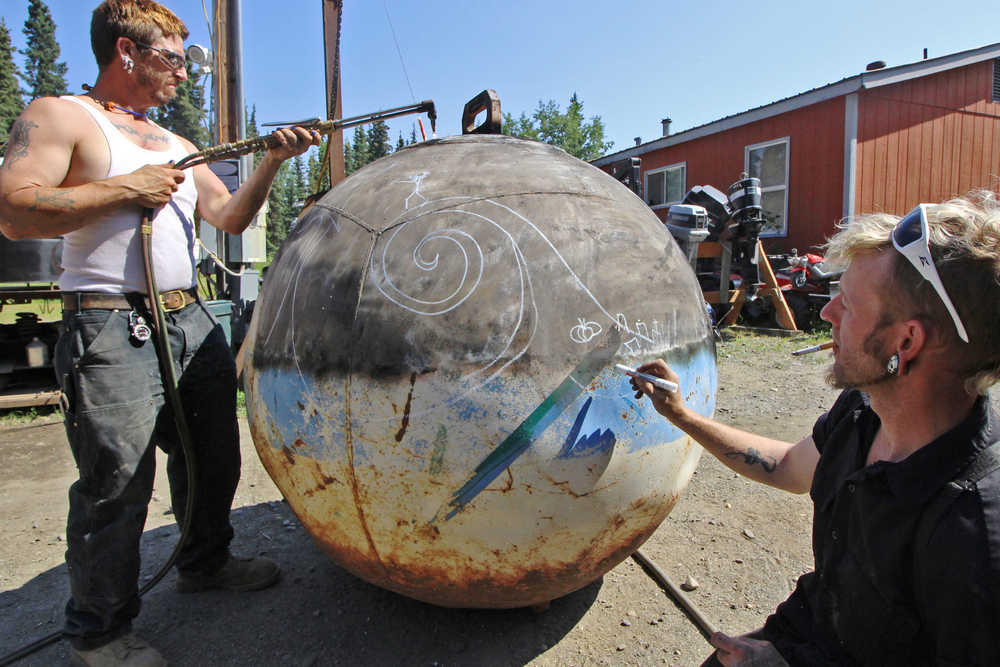 Ben Boettger/Peninsula Clarion RAW Welding employee Laron Hagan burns the paint off a steel buoy while owner Donnelle Scott sketches a "Nightmare Before Christmas"-inspired landscape at the RAW Welding workshop on Monday, July 18 outside Soldotna. The company will be plasma-cutting images into the buoy on Thursday at the Artzy Junkin craft market in Soldotna.