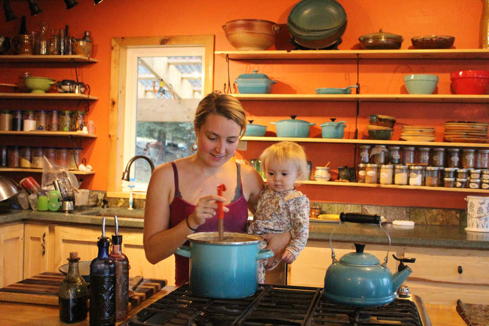 Eve Kilcher stirs a pot of chicken soup while holding Sparrow. The Kilcher's kitchen, which is the backdrop of the cover photo on Homestead Kitchen, was under construction for the majority of the time the Kilchers worked on the cookbook.