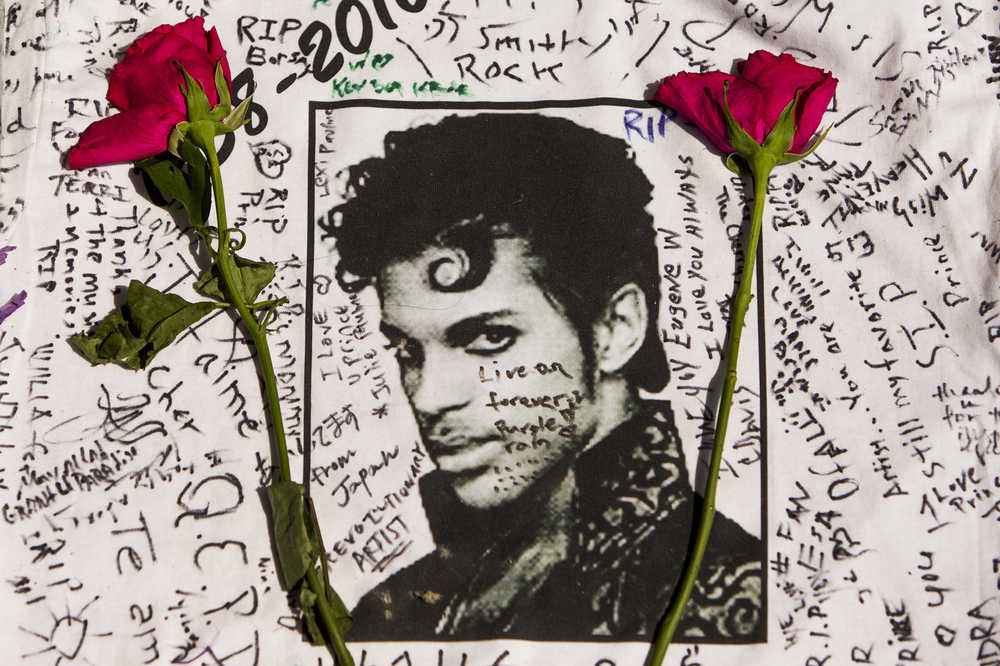 FILE - In this April 22, 2016 file photo, flowers lie on a T-shirt signed by fans at a makeshift memorial for musician Prince outside the Apollo Theater in New York. The singer died April 21, 2016, at the age of 57. With the loss of several icons of Generation X's youth, the year 2016 has left the generation born between the early 1960s and the early 1980s, wallowing in memories and contemplating its own mortality. (AP Photo/Andres Kudacki, File)