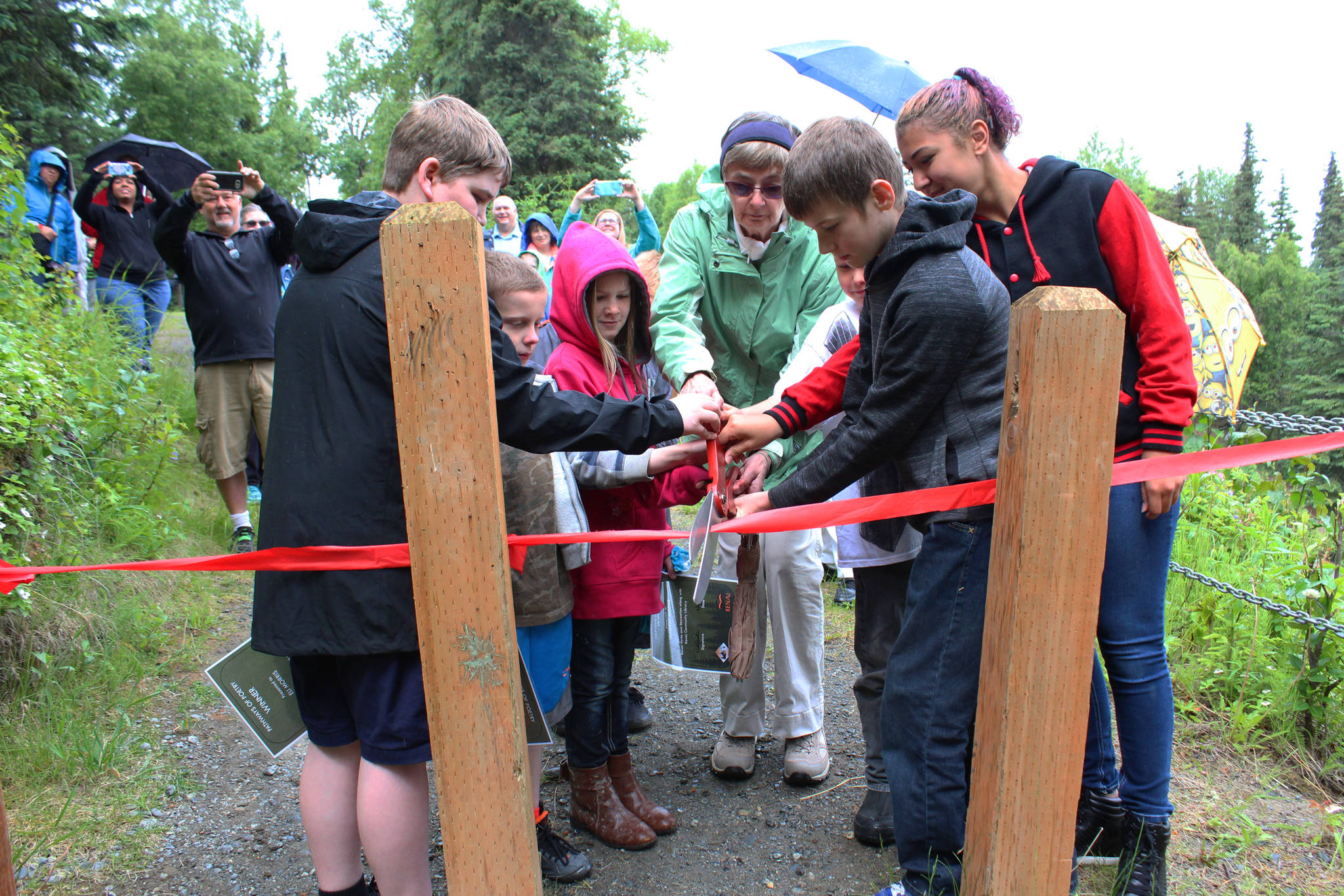 The winners of the Pathways of Poetry contest cut a ribbon opening a celebratory walk down the trail where their poems are displayed on signs Saturday, July 1, 2017 at Kenai Municipal Park in Kenai, Alaska. A panel of judges chose 12 winners out of 86 student participants, who submitted poems about nature. Scanning the codes on the signs brings up a recording of the authors reading their pieces. (Photo by Megan Pacer/Peninsula Clarion)