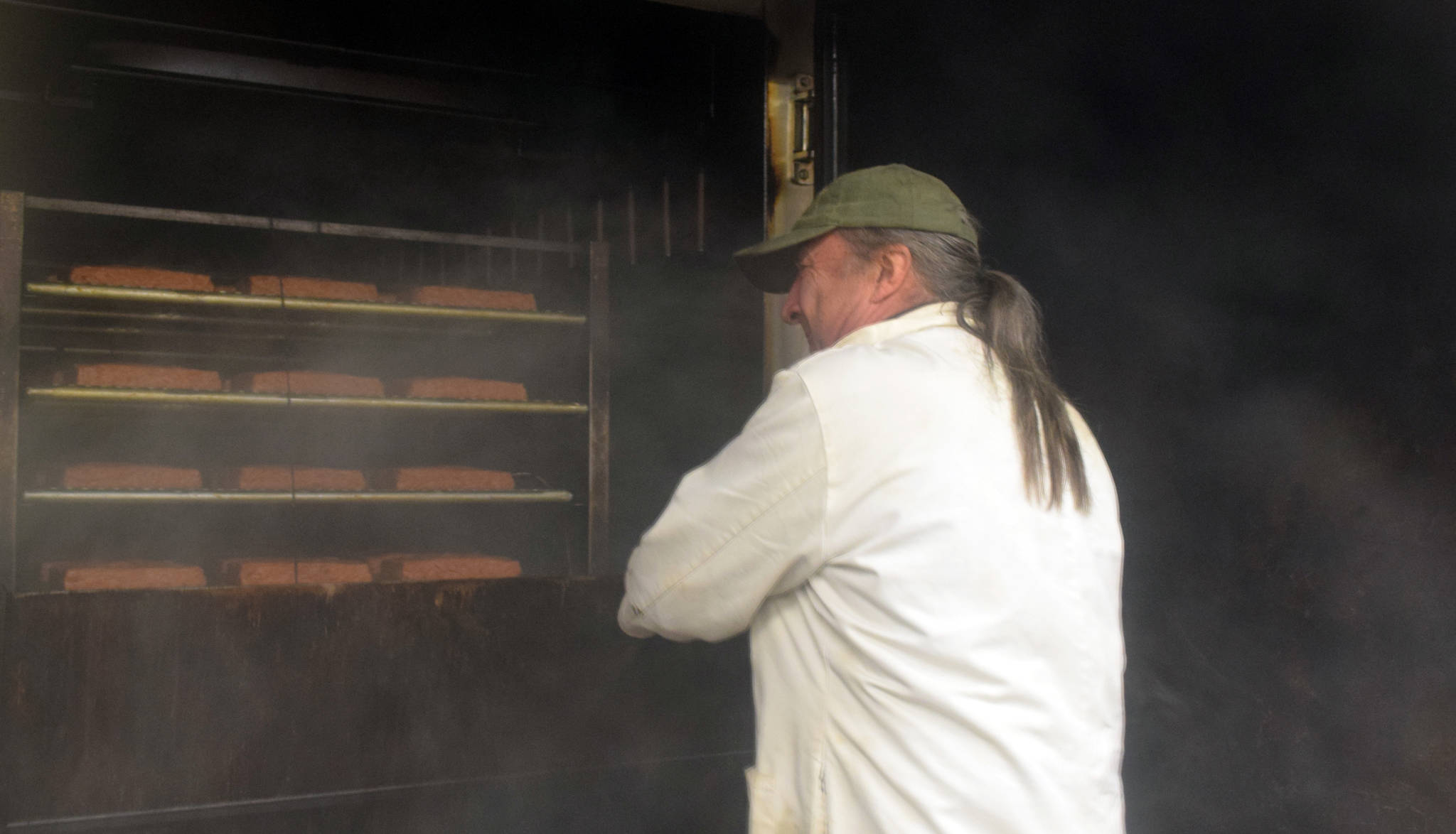 Through a haze of smoke, Fred West, owner of Tustumena Smokehouse, opens the door to the smoker where a batch of Kylee’s Alaska Salmon Bacon is smoking. (Photo by Kat Sorensen/Peninsula Clarion)