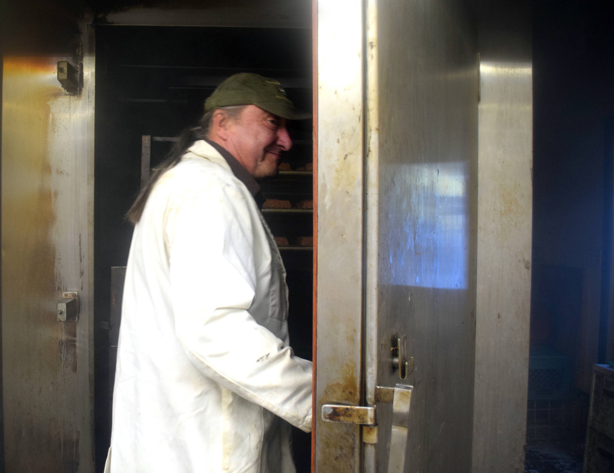 In a haze of smoke, Fred West, owner of Tustumena Smokehouse, opens the door to the smoker where a batch of Kylee’s Alaska Salmon Bacon is smoking. (Photo by Kat Sorensen/Peninsula Clarion)