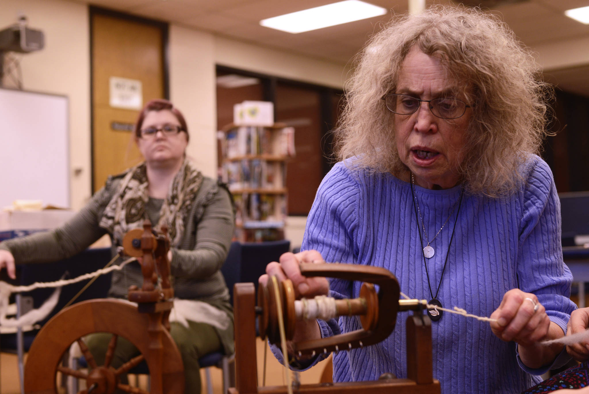 Instructor Lee Coray-Ludden (center) shows how to spin yarn from wool on a kick-driven spinning wheel, with student Amy Townson practicing in the background on Tuesday, March 20, 2018 in the Soldotna Preparatory School library in Soldotna, Alaska. In her four-session weekly yarn spinning class, part of the Soldotna Community Schools program, Coray-Ludden will teach students to spin and ply yarn from locally-grown wool and fleece, including exotic fibers from cashmere goats, alpacas, and llamas. (Photo by Ben Boettger/Peninsula Clarion).