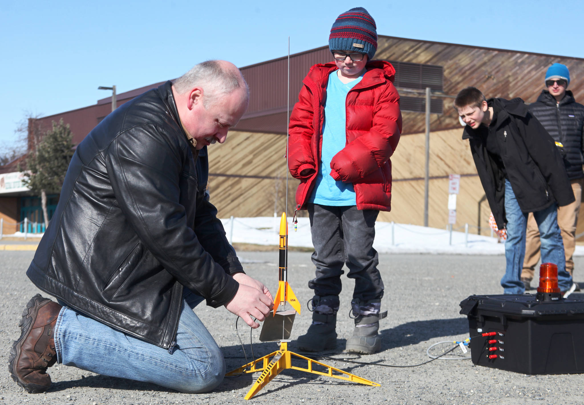 Scott Aleckson (left) clips wires to the ignition contacts of a model rocket while students at his model rocketry session observe in the parking lot of the Soldotna Regional Sports Complex in Soldotna, Alaska on Saturday, May 31, 2018. Aleckson said he made and launched his first plastic, balsa wood, and cardboard rocket as a 4th grade student at Soldotna Elementary School. Since then he’s built a custom launch system, he said, “with recycled electronics and a lot of soldering time.” Aleckson taught Saturday’s rocketry class through the Soldotna Community Schools program, and said he’d like to continue spreading the hobby. “If I could get a model rocket club going, that’d be great,” he said. “Stuff like this is much more fun in a group.” (Photo by Ben Boettger/Peninsula Clarion)