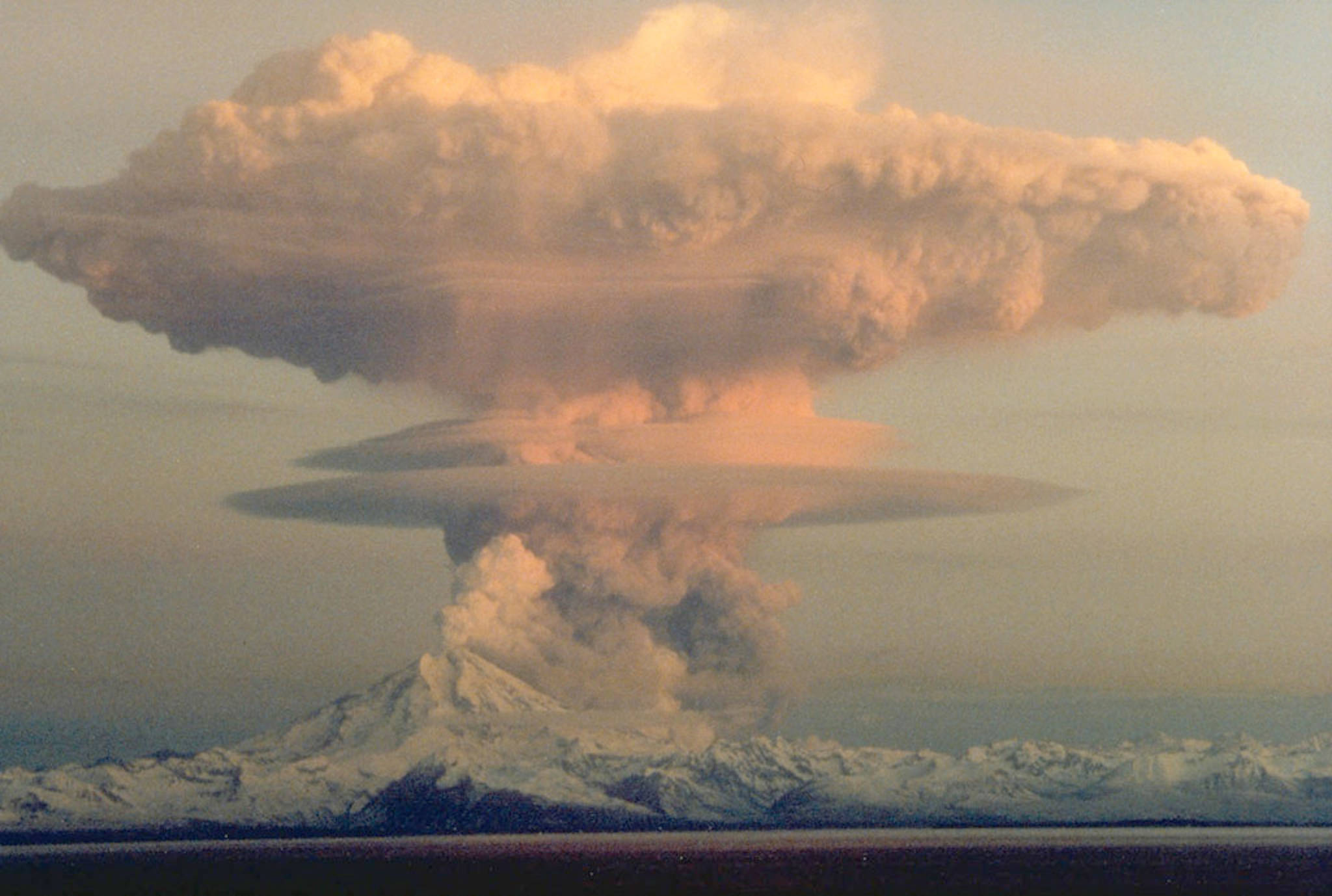 This April 21, 1990 image shows an ascending eruption cloud from Redoubt Volcano as viewed to the west from the Kenai Peninsula, Alaska. The mushroom-shaped plume rose from avalanches of hot debris that cascaded down the north flank of the volcano. A smaller, white steam plume rises from the summit crater. (Image courtesy Robert Clucas and the U.S. Geological Survey)