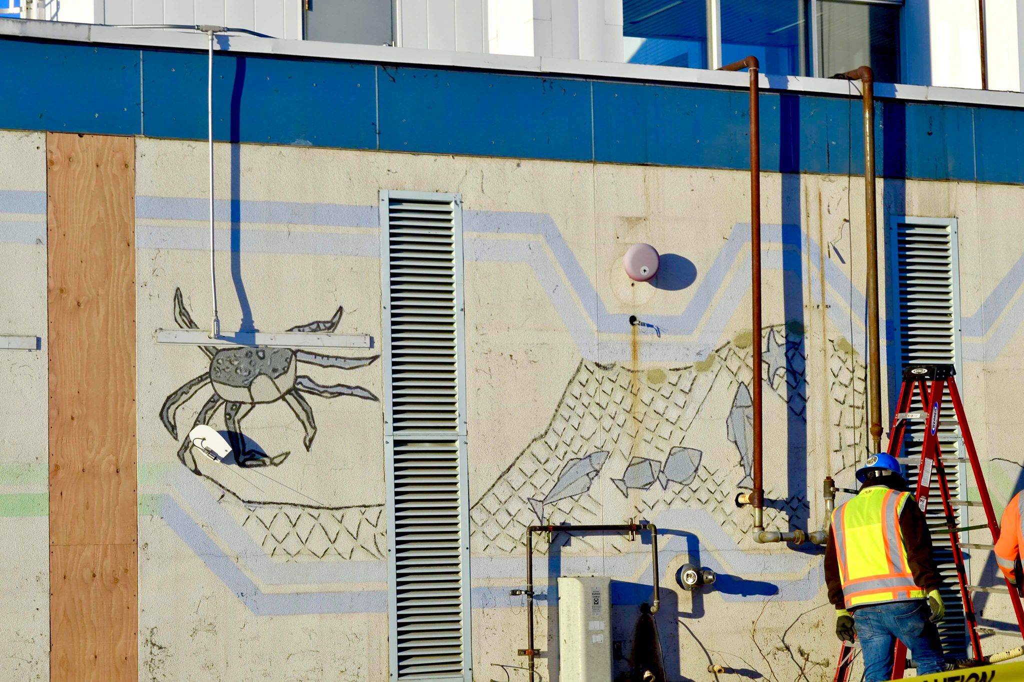 Construction crews work at the Kenai Municipal Airport on Tuesday afternoon. After workers removed siding from the facade, a mural with iconic Kenai images like fishing nets, the St. Nicholas Russian Orthodox chapel and a dog musher, was revealed. (Photo by Victoria Petersen/Peninsula Clarion)