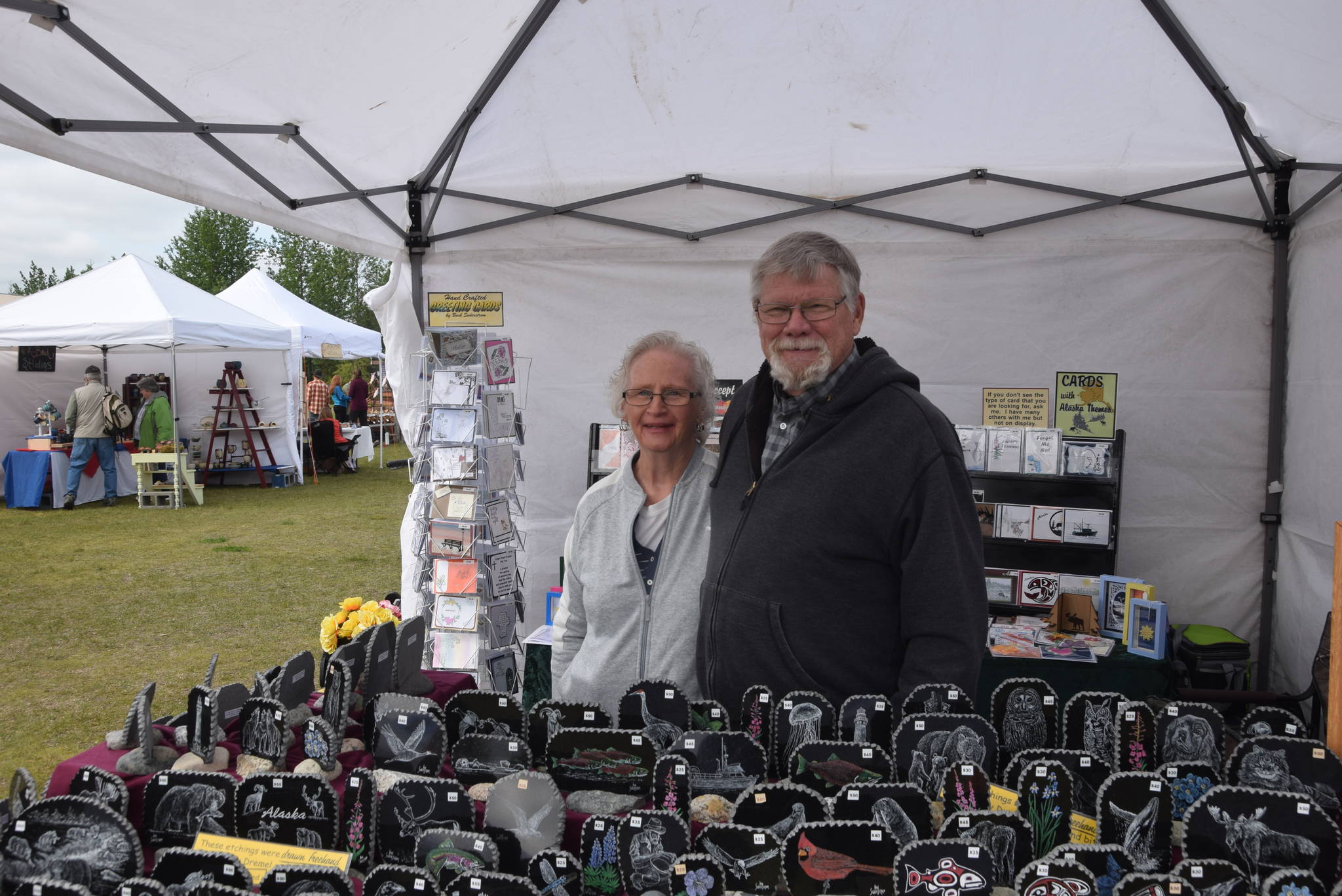 Barb and Don Soderstrom pose in front of their artwork, handmade greeting cards by Barb and etchings in granite by Don, during the Kenai River Festival at Soldotna Creek Park in Soldotna, Alaska on June 8, 2019. (Photo by Brian Mazurek/Peninsula Clarion)