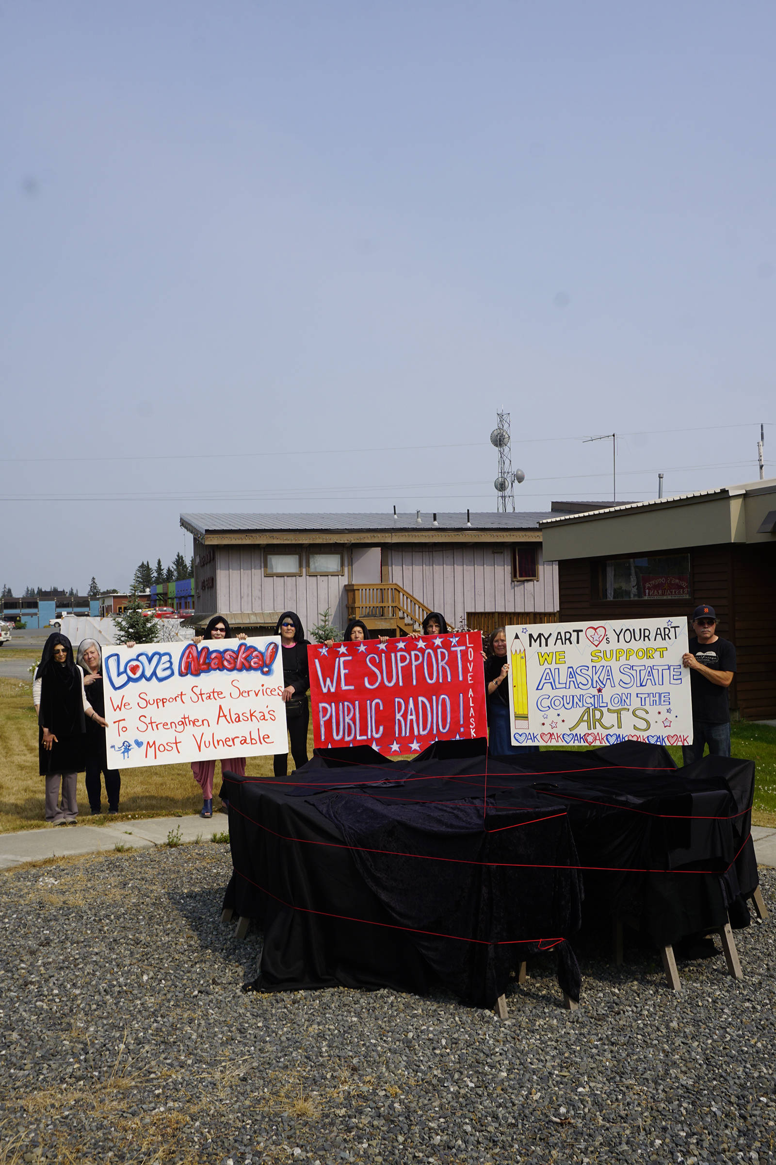 Arts supporters draped in black holds sings on July 9, 2019, by Sean Derry’s public art sculpture in Homer, Alaska, as part of a statewide art intervention to protest Gov. Mike Dunleavy’s veto of a $2.8 million state appropriation to the Alaska State Council on the Arts. They also supported a general override of Dunleavy’s vetoes that will affect funding for the University of Alaska, public radio and other programs. Derry’s sculpture was commissioned as a 1% for art project associated with the remodeling of Pioneer Hall at the Kachemak Bay Campus, Kenai Peninsula College, University of Alaska. The protest was not sanctioned by the college. (Photo by Michael Armstrong/Homer News)