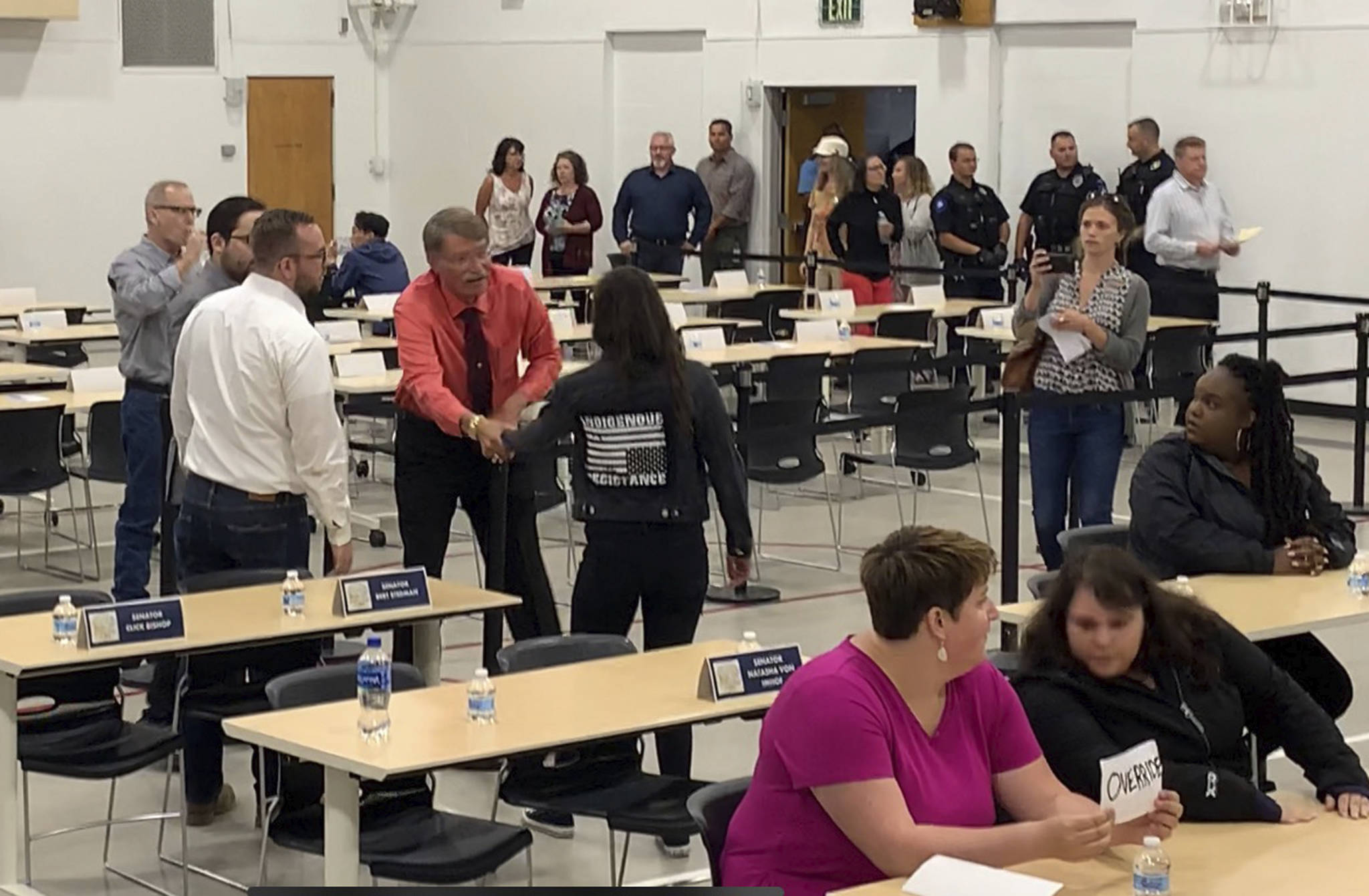 In this July 10, 2019, photo provided by Rochelle Adams, the mayor of Wasilla, Alaska, Bert Cottle, center left in the orange shirt, is seen grabbing the arm of Alaska Native activist Haliehana Stepetin, who was trying to take part in a sit-in of legislators meeting in Wasilla. Stepetin filed an assault complaint with the Wasilla Police Department on Thursday, naming Cottle and Zachary Freeman, the spokesman for the Republican House Minority, seen next to Cottle in the white dress shirt. (Rochelle Adams via AP)
