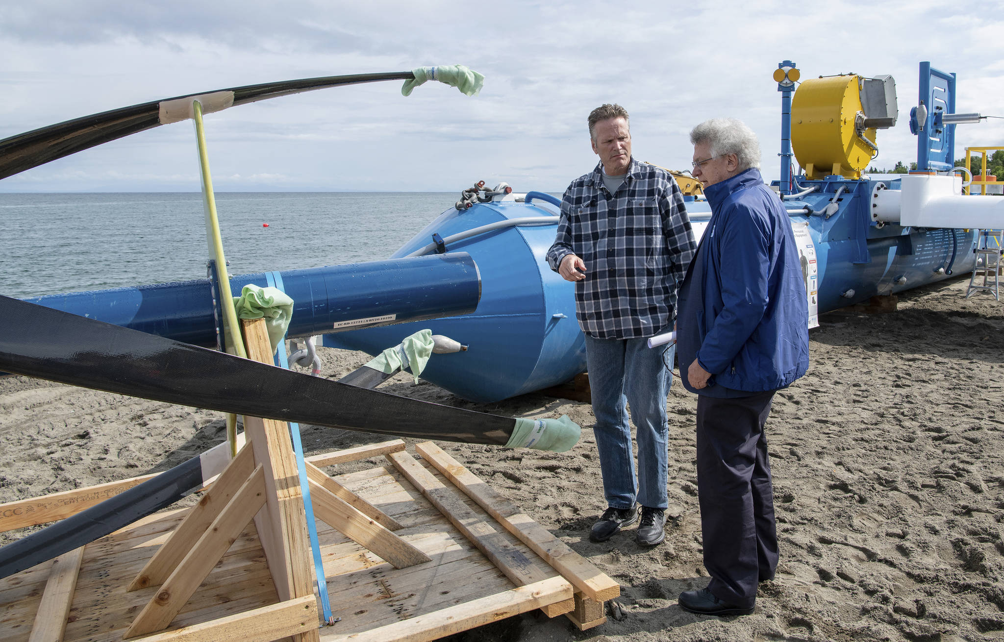 In this Tuesday, July 16, 2019, photo released by the Alaska Governor’s office, Gov. Mike Dunleavy, left, talks with ORPC CEO, Co-Founder, and Chairman Chris Sauer in front of a Riv-Gen Power System turbine on the bank of the Kvichak River in Igiugig, Alaska. A tiny Alaska Native village is adopting an emerging technology to transform the power of a local river into a renewable energy source. (Austin McDaniel/Alaska Governor’s Office via AP)
