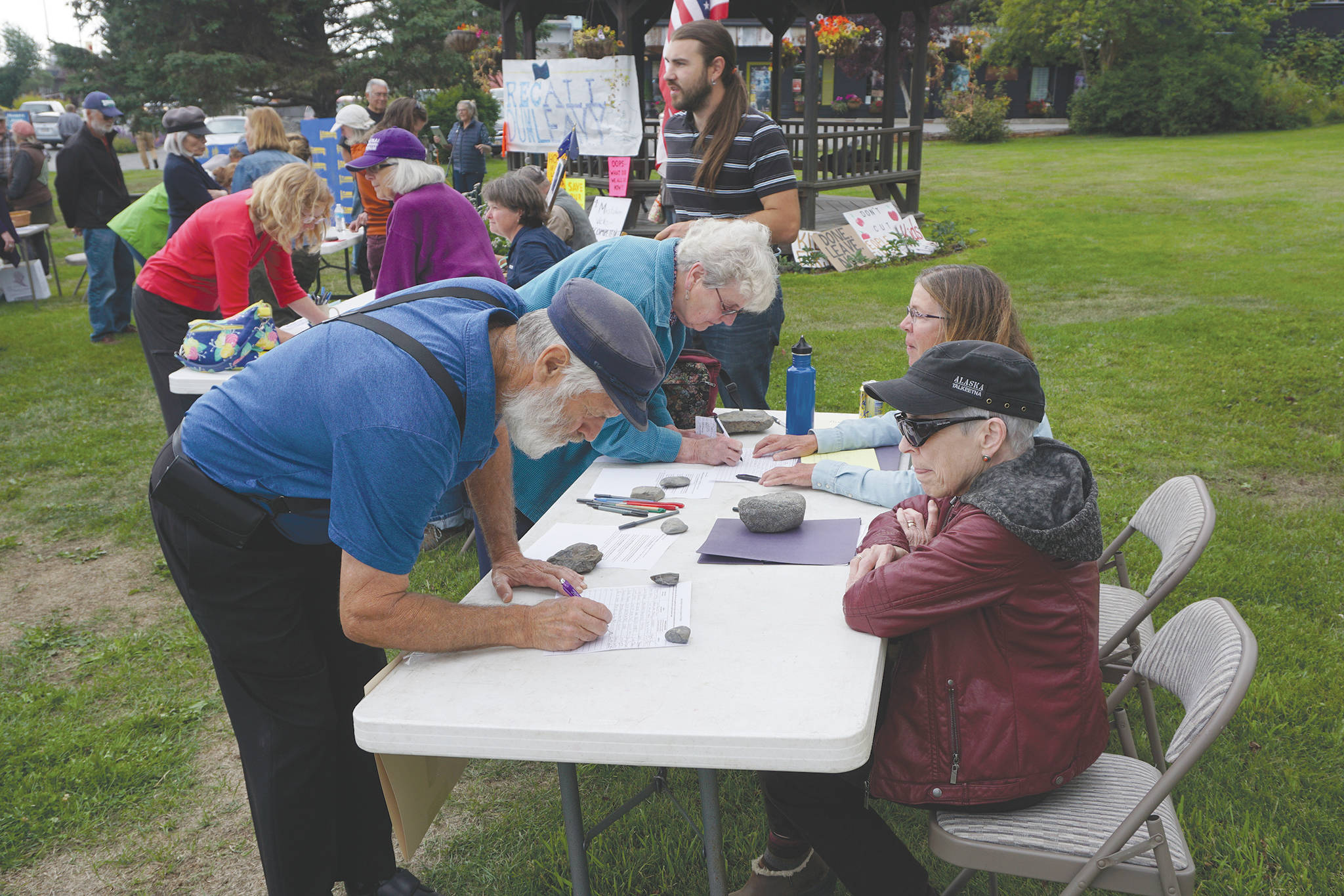 Recall Dunleavy organizers Ann Keffer, right, in hat, and Pat Cue, behind Keffer, take signatures at a Recall Dunleavy rally held on Aug. 1 at WKFL Park in Homer. At left, former Homer Rep. Paul Seaton, NP-Homer, signs a form. Seaton lost to Rep. Sarah Vance, R-Homer, in the general election. (Photo by Michael Armstrong)