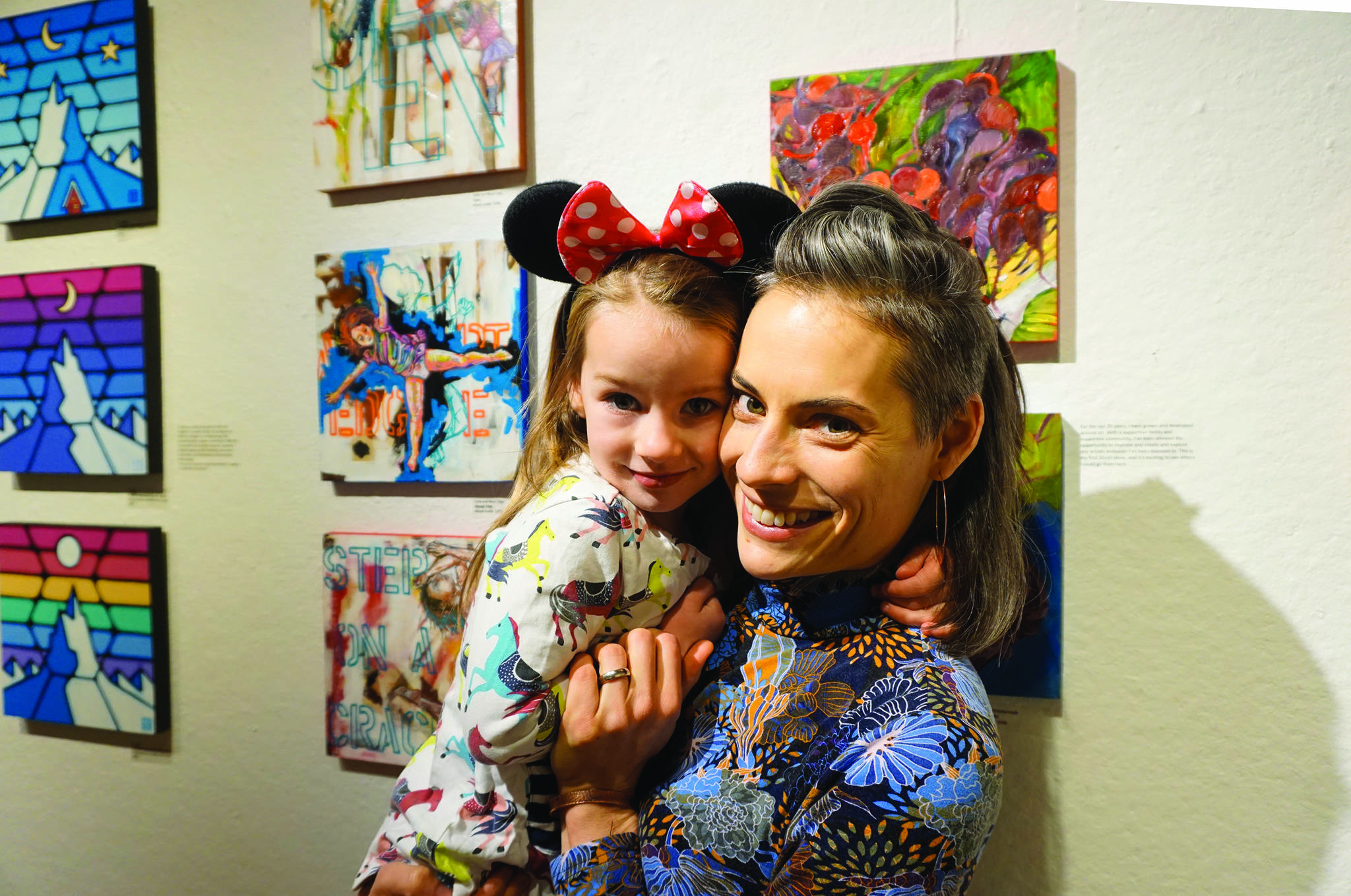 Carla Cope and her daughter, Nova, pose next to their paintings at left at the Nov. 8, 2019, opening of the 10x10 show at Bunnell Street Arts Center in Homer, Alaska. Nova painted the background elements to the paintings and her mother added to that. (Photo by Michael Armstrong/Homer News)