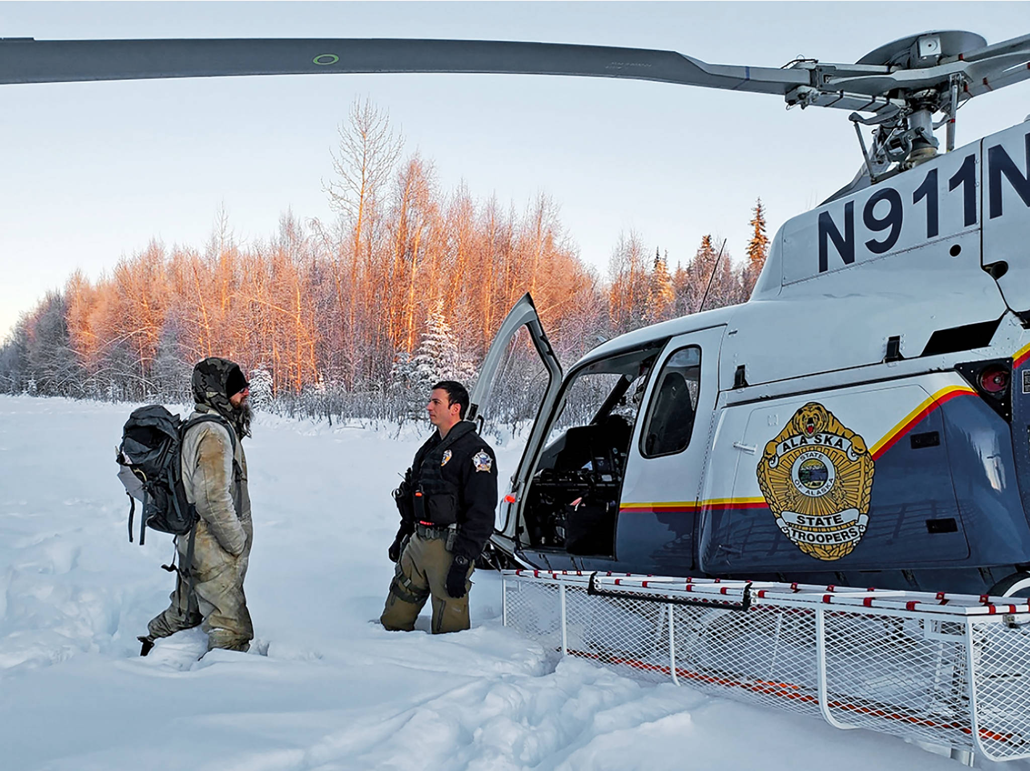 This Jan. 9, 2020 photo provided by Alaska State Troopers shows Tyson Steele, left, talking with Helo 3 Tactical Flight Officer Zac Johnson after Steele’s rescue outside Susitna Valley, Alaska. Troopers rescued Tyson Steele, 30, who survived in a makeshift shelter after his remote cabin burned last month, after spotting him and an SOS sign in the snow. (Alaska State Troopers via AP)