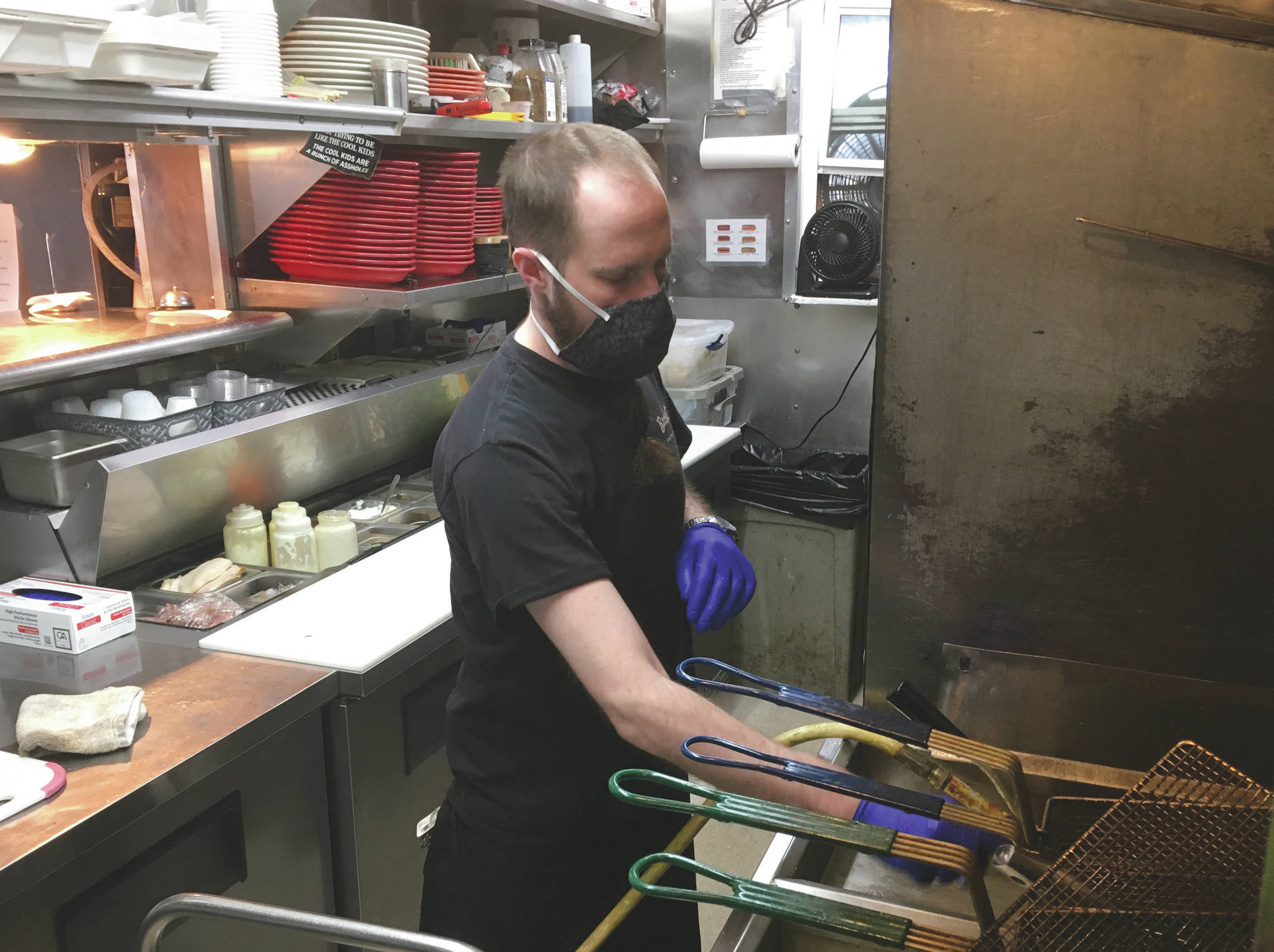 Trevor Pound, a line cook at Buckets Sports Grill in Soldotna, filters the friers Thursday, April 30, 2020. Despite the heat in the kitchen, state regulations say all restaurant employees must wear cloth face masks. “It’s completely miserable, dude,” Pound said. “It gets so hot back here I can’t breathe without a face mask. But I’ve got to pay the bills, so I wear a mask.” (Photo by Jeff Helminiak/Peninsula Clarion)