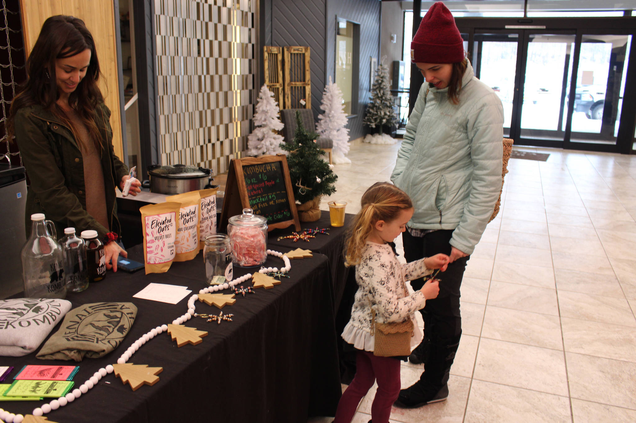 Remmy Rutledge, center, and her mom Breanna Rutledge, right, of Sterling, check out Remmy’s new ornament purchased from Kenai Kombucha owner Devon Gonzales, left, during the Merry Little Christmas Market at the Peninsula Center Mall in Soldotna, Alaska, on Nov. 7, 2020. (Photo by Brian Mazurek/Peninsula Clarion)