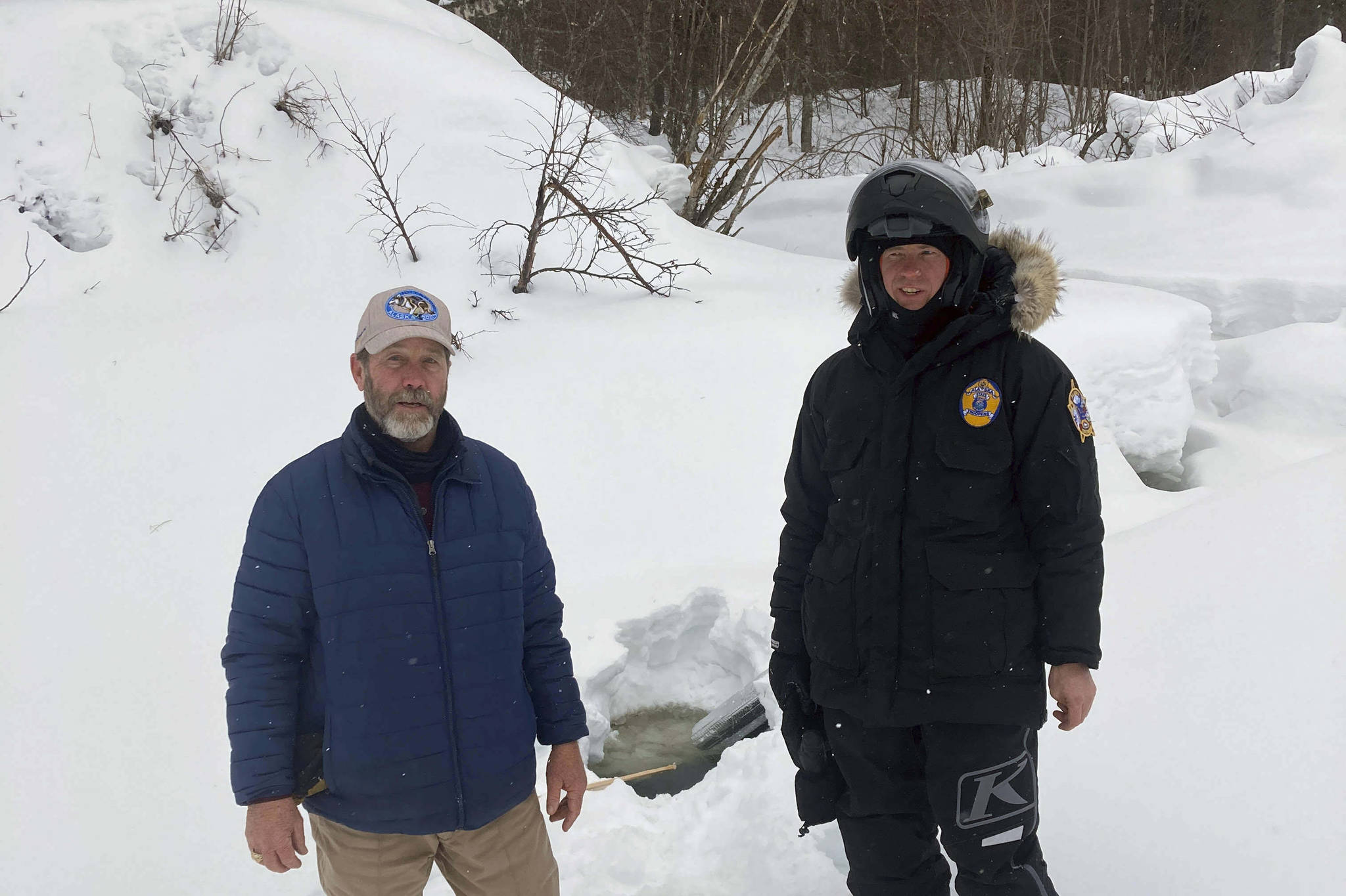 This photo provided by the Alaska Wildlife Troopers taken March 9, 2021, Doug Ramsey, left, of Sundance Wyoming, poses with Alaska Wildlife Trooper Jason Kneier near a hole in the ice of a river in Swentna, Alaska. The two helped pull an 8-year-old boy from the water after he fell into the river. (Alaska Wildlife Troopers via AP)