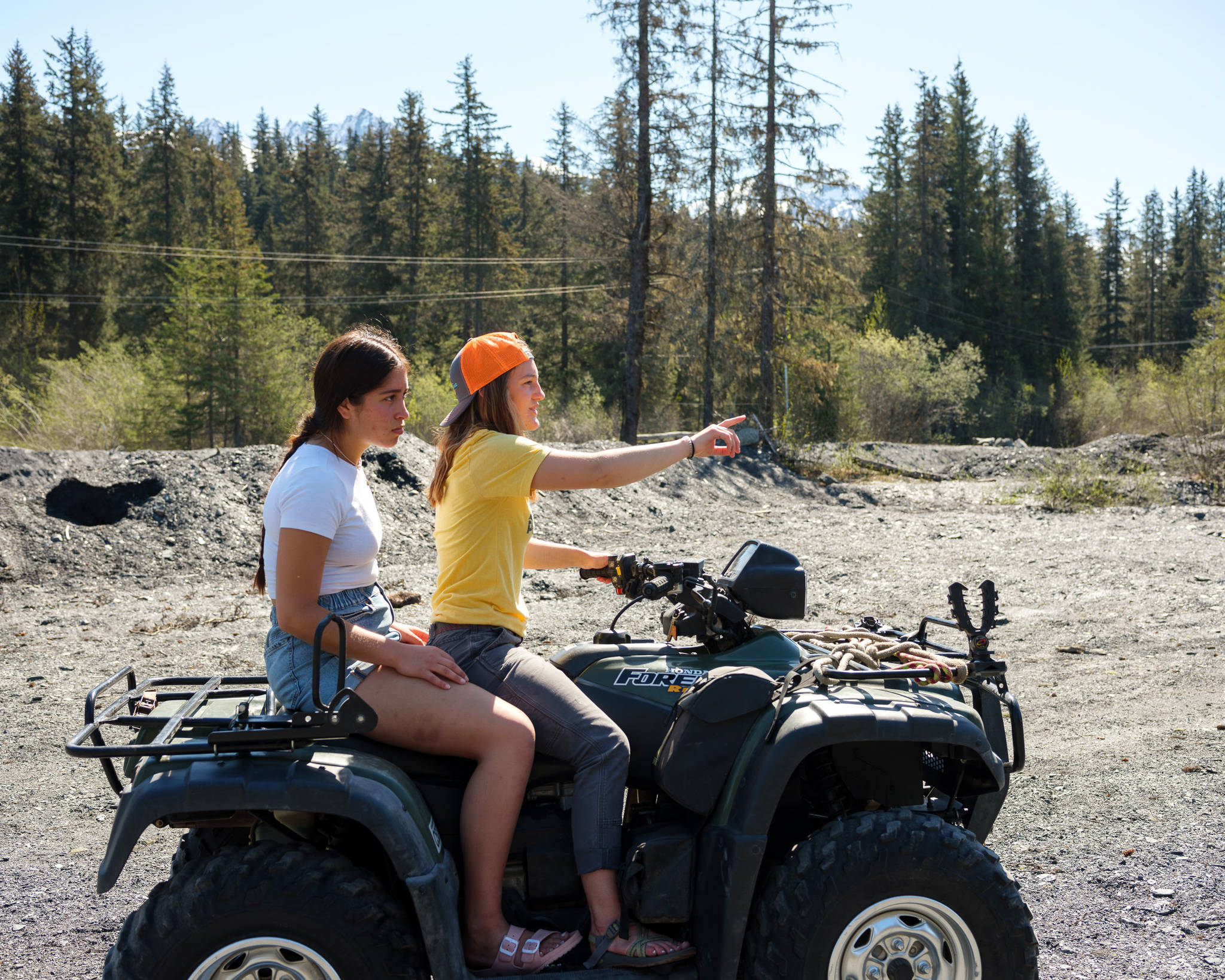 In May 2021, Selma Casagranda, 18, and Annika Nilsson, 17, ride the Nilsson family ATV along Lost Creek — the stream that floods Nilsson’s neighborhood in Seward, Alaska, each fall — and point to where a house once stood before flood waters made the property useless. (Young Kim for The Hechinger Report)