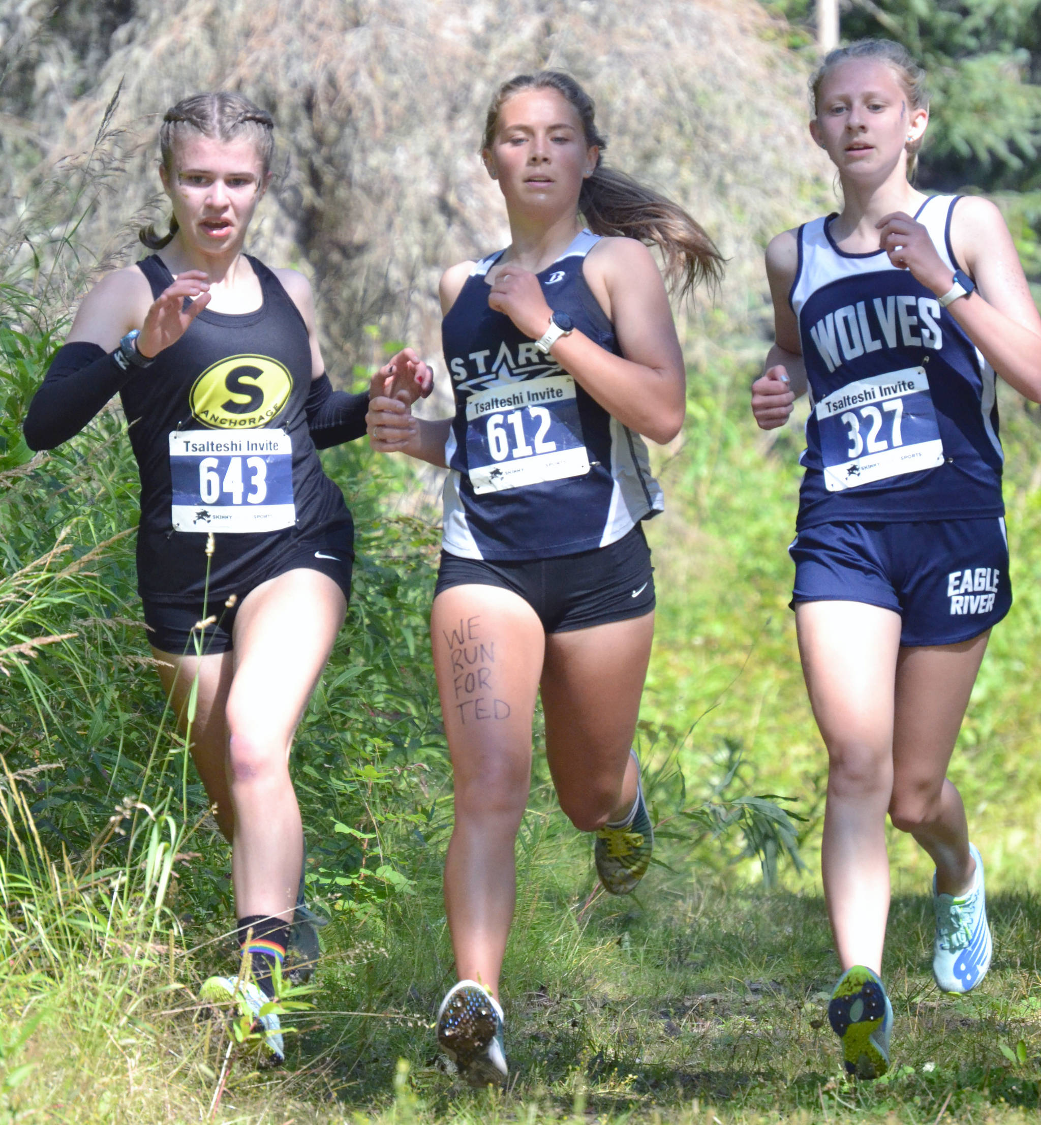 Soldotna’s Jordan Strausbaugh races to 17th place in the girls varsity race at the Ted McKenney XC Invitational on Saturday, Aug. 21, 2021, at Tsalteshi Trails just outside of Soldotna, Alaska. “We run for Ted” is written on Strausbaugh’s thigh in honor of former Soldotna coach Ted McKenney. Strausbaugh runs with South’s Elizabeth Page and Eagle River’s Ava Trembath. (Photo by Jeff Helminiak/Peninsula Clarion)