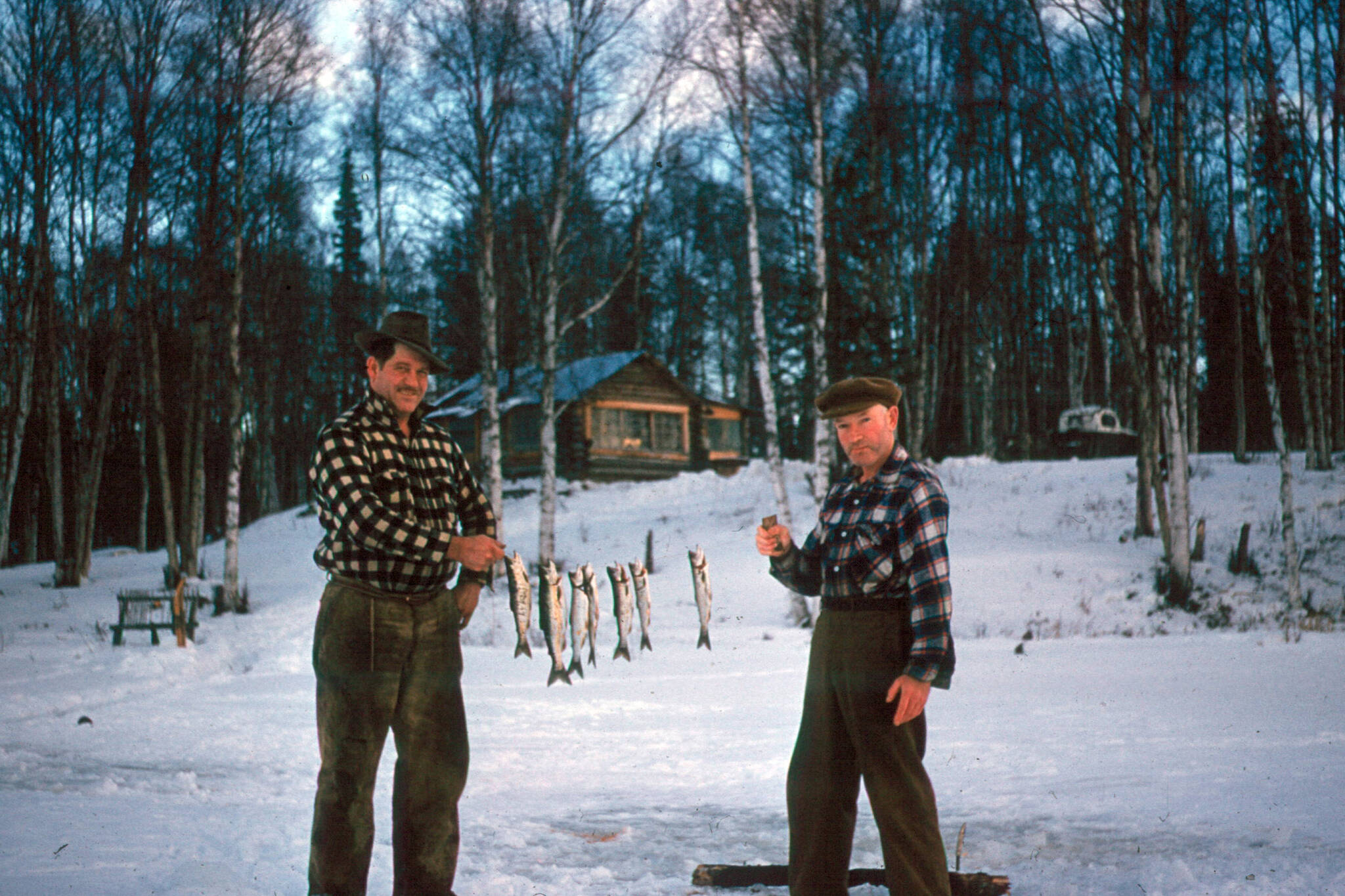 In this 1950s image, Chell Bear (left) and Lawrence McGuire display a stringer of small trout they caught through the ice in front of the homestead cabin of Bob Mackey, for whom the Mackey Lakes were named. (Photo courtesy of the Kenai Peninsula College Historic Photo Repository)