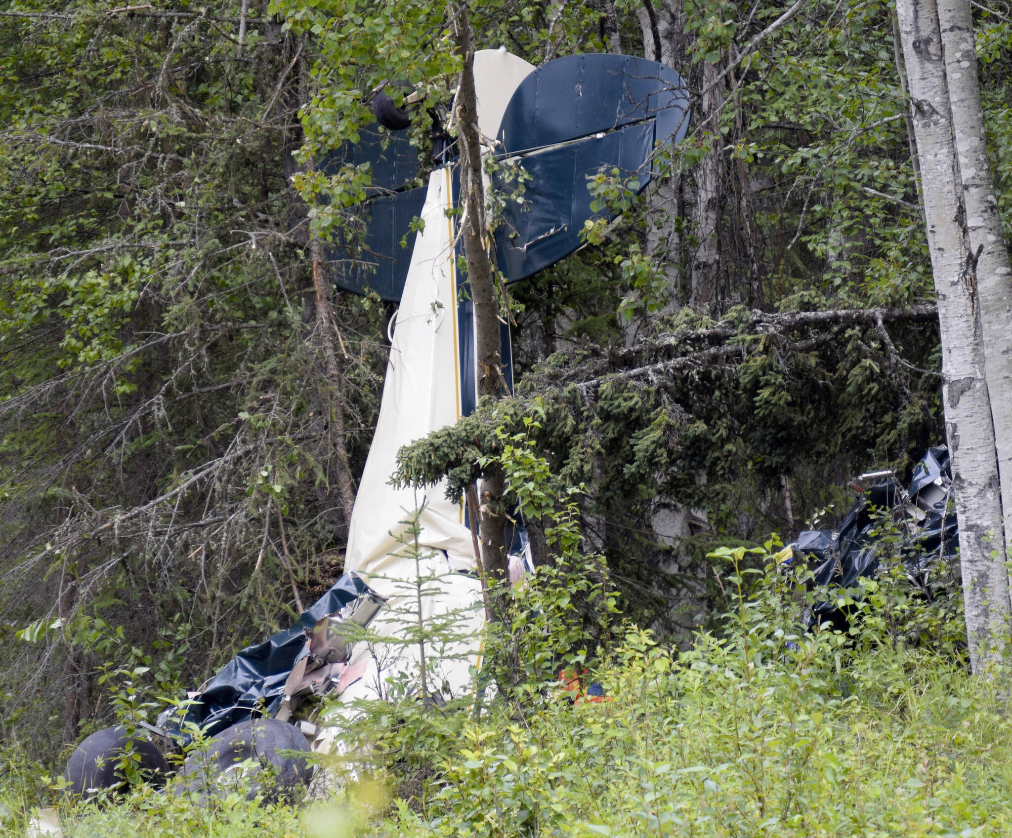 In this July 31, 2020, file photo, a plane rests in brush and trees outside of Soldotna, Alaska, after a midair collision that killed seven people, including State Rep. Gary Knopp. A Feb. 22 report by the National Transportation and Safety Board is recommending that all pilots be required to communicate their positions on a designated radio frequency when entering and exiting areas not controlled by air traffic control towers throughout Alaska. (Jeff Helminiak/Peninsula Clarion via AP, F-le)