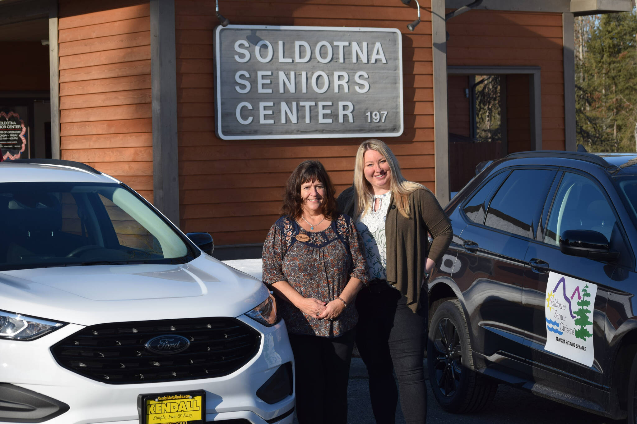 Loretta Knudson-Spalding, left, and Regional Director of the Office of the Governor Jill Schaefer, right, pose next to the Soldotna Senior Center’s new cars in Soldotna on Wednesday, March 30, 2022. (Camille Botello/Peninsula Clarion)