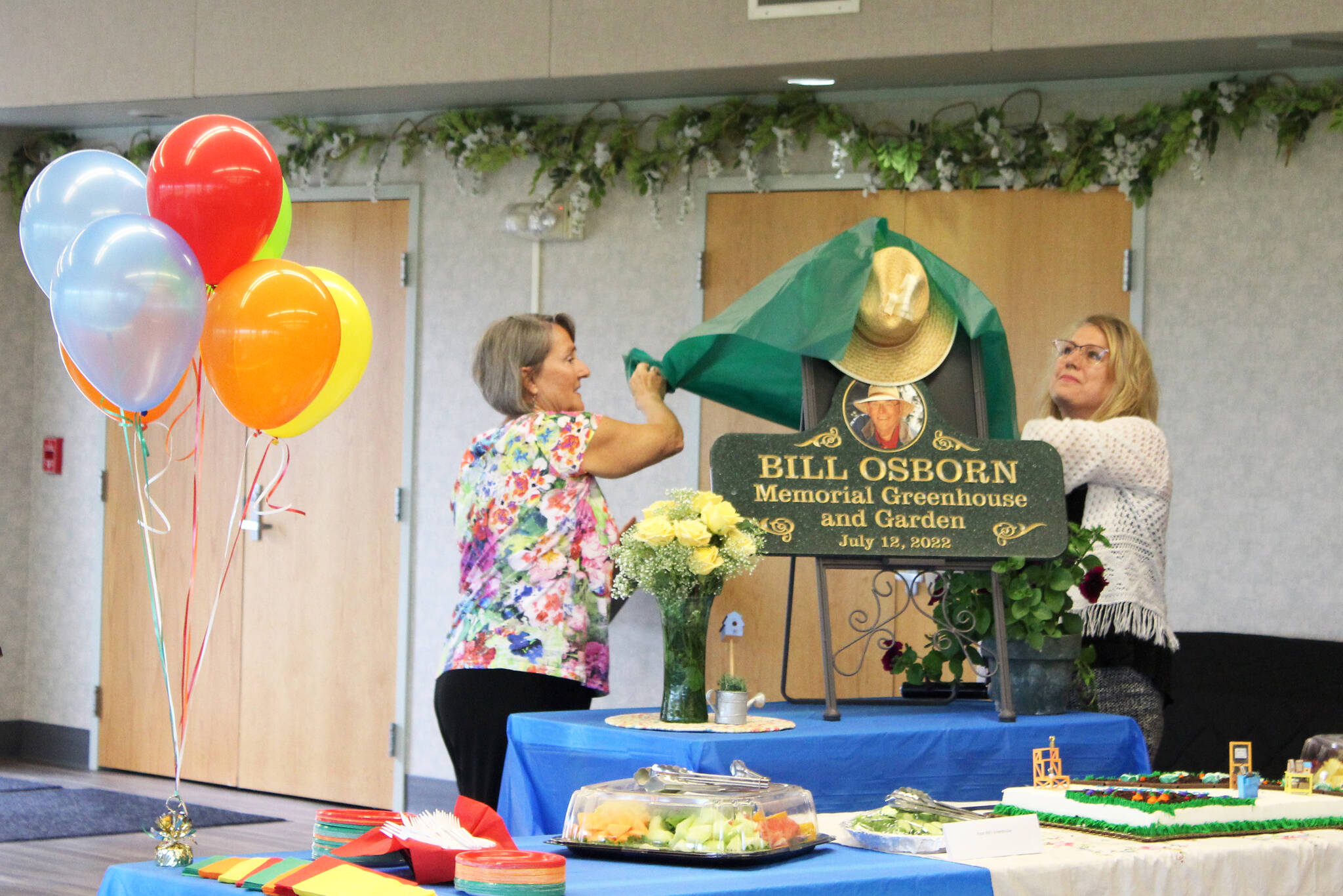 Vicke Kukowski (left) and Kathy Romain (right) unveil a plaque dedicating the Kenai Senior Center’s greenhouse to Bill Osborn during a ceremony on Tuesday, July 12, 2022 in Kenai, Alaska. (Ashlyn O’Hara/Peninsula Clarion)