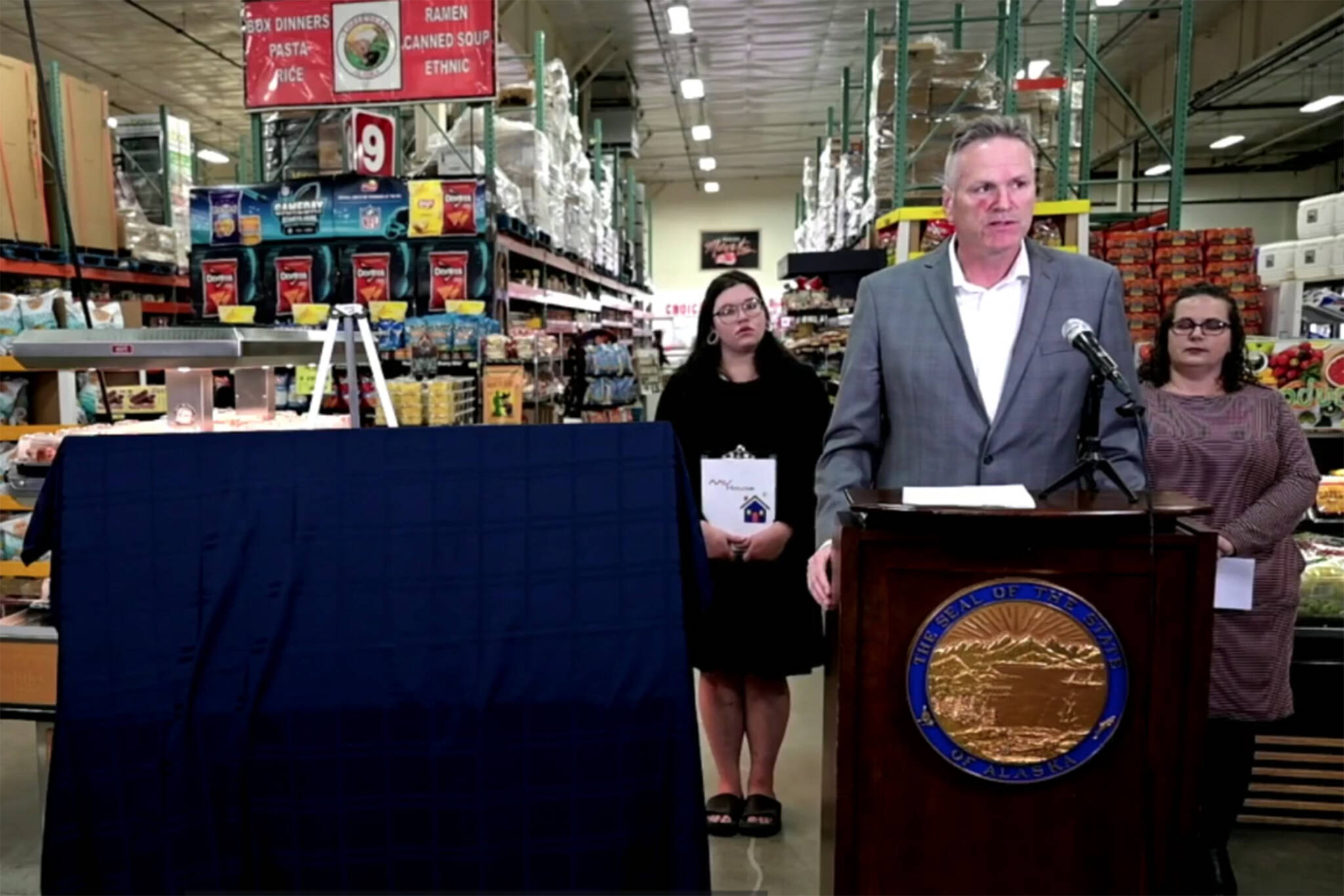 Gov. Mike Dunleavy stands at a lectern in a Three Bears Alaska store in Palmer, next to a giant, covered check containing this year’s Alaska Permanent Fund dividend amount on Thursday, Sept. 8, 2022. Behind him stand Alaska resident Miranda Wagoner, left, and Jessica Viera, executive director, Wasilla Chamber of Commerce, who gave speeches at the event. (Faceboook live screenshot)