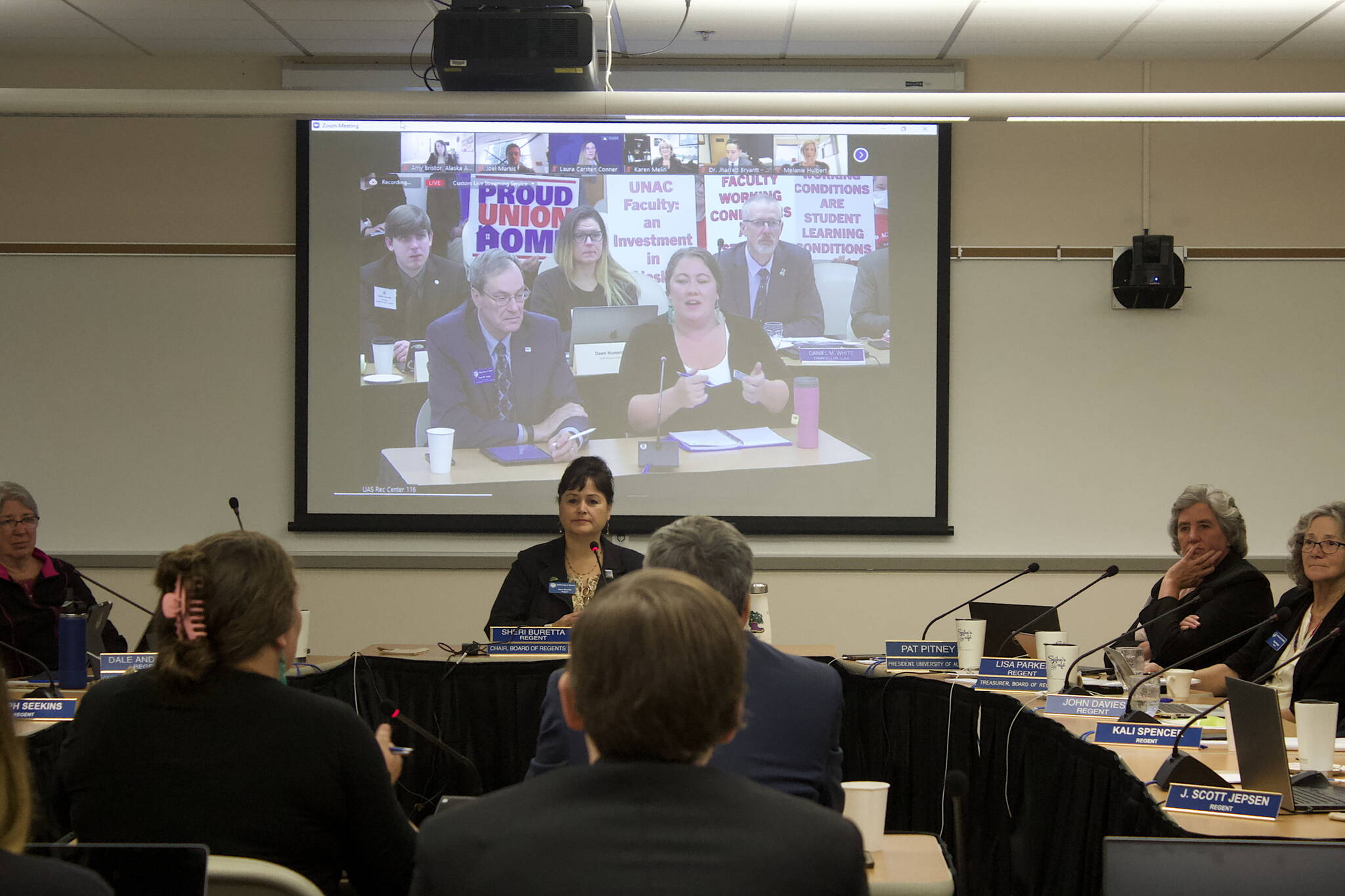 Amanda Triplett, at right on overhead screen, discusses the University of Alaska’s “Did You Know” program while faculty members in the background hold up signs protesting a labor contract stalemate during the Board of Regent’s meeting Friday in Juneau. The program highlights cooperative efforts the university is participating in with communities such as workplace experience and dual enrollments with other educational institutions. (Mark Sabbatini / Juneau Empire)