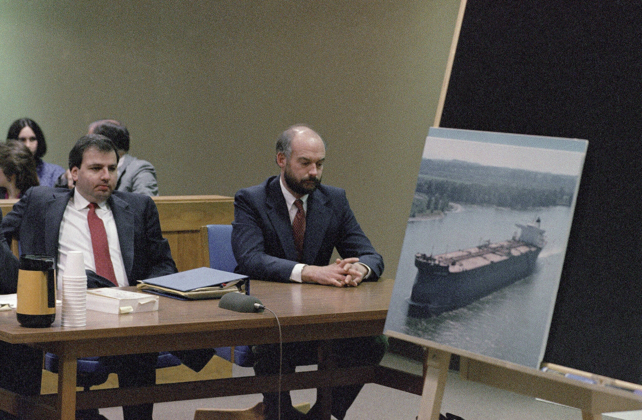Fired Exxon Valdez skipper Joseph Hazelwood, right, sits with his lawyer Michael Chalos as a photograph of the tanker is displayed on an easel in Anchorage Superior Court, March 20, 1990. Hazelwood, the captain of the Exxon Valdez oil tanker that ran aground more than three decades ago in Alaska, causing one of the worst oil spills in U.S. history, has died in July 2022, the New York Times reported. He was 75. (AP Photo/Jack Smith, File)