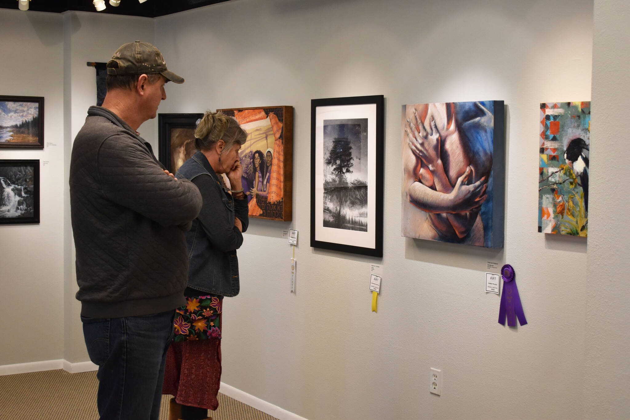 Attendees of the opening reception of the Kenai Art Center Biennial Juried Show look at a wall of art that includes Best of Show winner “Cross My Heart,” an untitled photograph which won honorable mention, the third place winner “Civil Disobedience,” and Juror’s Choice “The Dead Skate,” on Thursday, Oct. 6, 2022, at the Kenai Art Center in Kenai, Alaska. (Jake Dye/Peninsula Clarion)