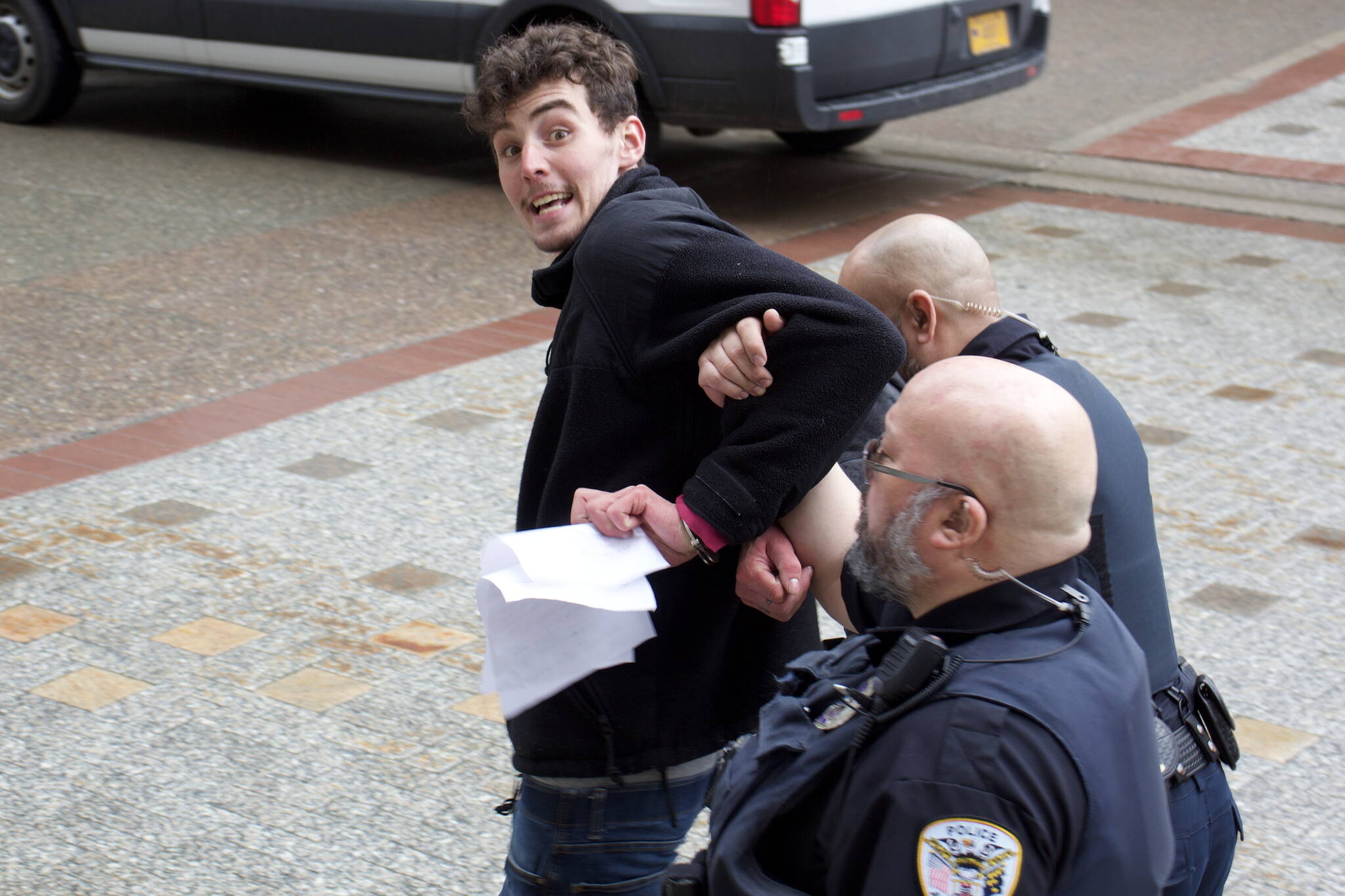 Eric Osuch tries to offer papers related to his arrest in front of the Alaska State Capitol on Monday, April 17, 2023, to a reporter as Juneau Police Department officers escort him to a nearby patrol vehicle. Osuch, who was staging a solo protest about fisheries bycatch policies, was banned from the Capitol after causing a public disruption and was arrested a short time later for another alleged disturbance inside the State Office Building. (Mark Sabbatini / Juneau Empire)