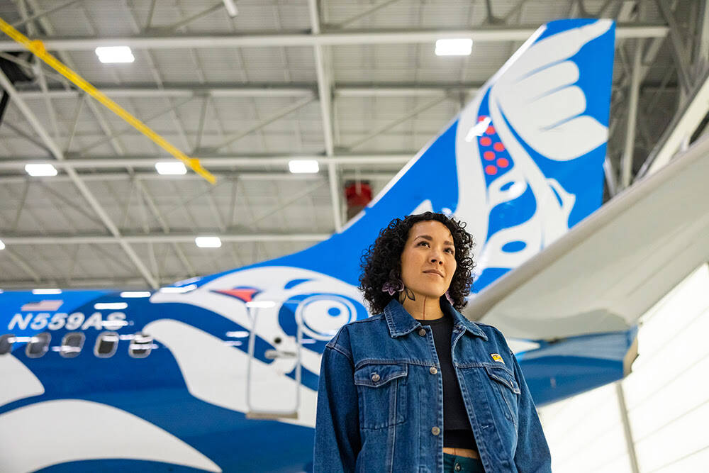 Juneau artist Crystal Kaakeeyáa Worl poses with an Alaska Airlines 737-800 aircraft decorated with Worl’s latest work, Xáat Kwáani (Salmon People). Alaska Airlines held a unveiling ceremony on Friday, May 12 to welcome the plane into service. (Courtesy Photo / Alaska Airlines)