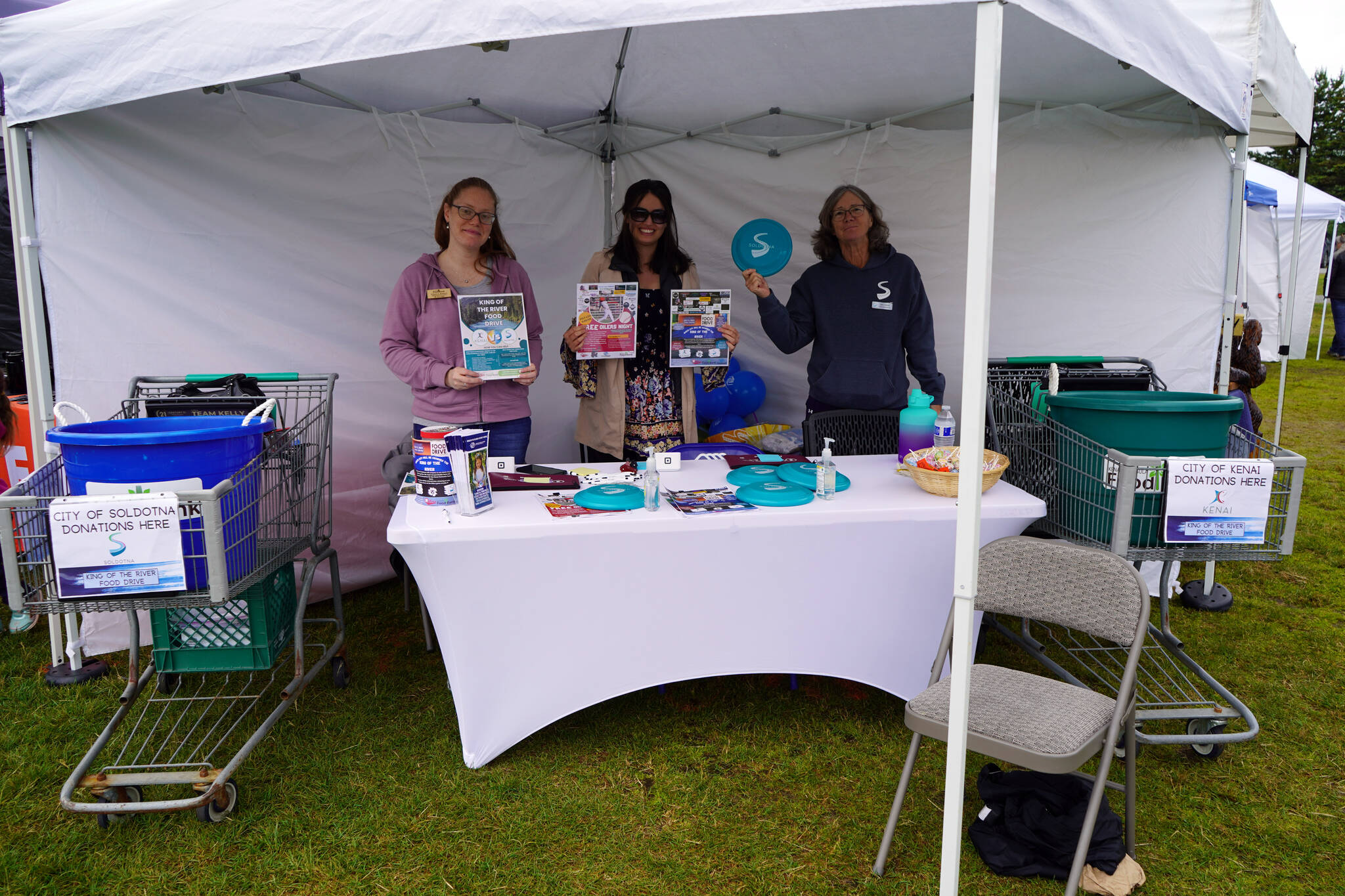Kenai Peninsula Food Bank Administrative Assistant Claire Jones and Donations and Communications Manager Lilly Murray stand with Soldotna Vice Mayor Lisa Parker as they fundraise for the King of the River Food Drive at the Wednesday Market in Soldotna Creek Park in Soldotna, Alaska, on Wednesday, June 28, 2023. (Jake Dye/Peninsula Clarion)