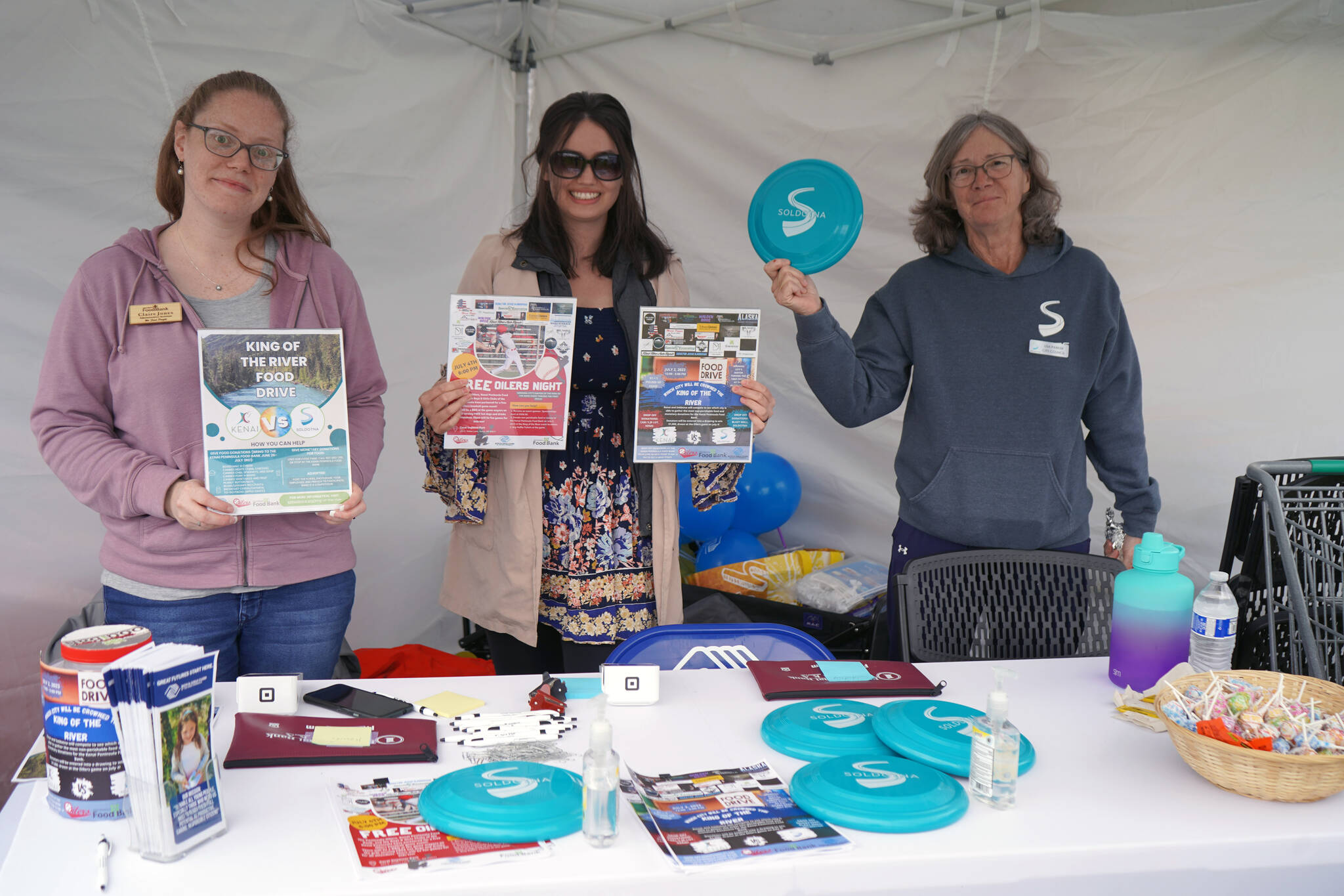 Kenai Peninsula Food Bank Administrative Assistant Claire Jones and Donations and Communications Manager Lilly Murray stand with Soldotna Vice Mayor Lisa Parker as they fundraise for the King of the River Food Drive at the Wednesday Market in Soldotna Creek Park in Soldotna, Alaska, on Wednesday, June 28, 2023. (Jake Dye/Peninsula Clarion)