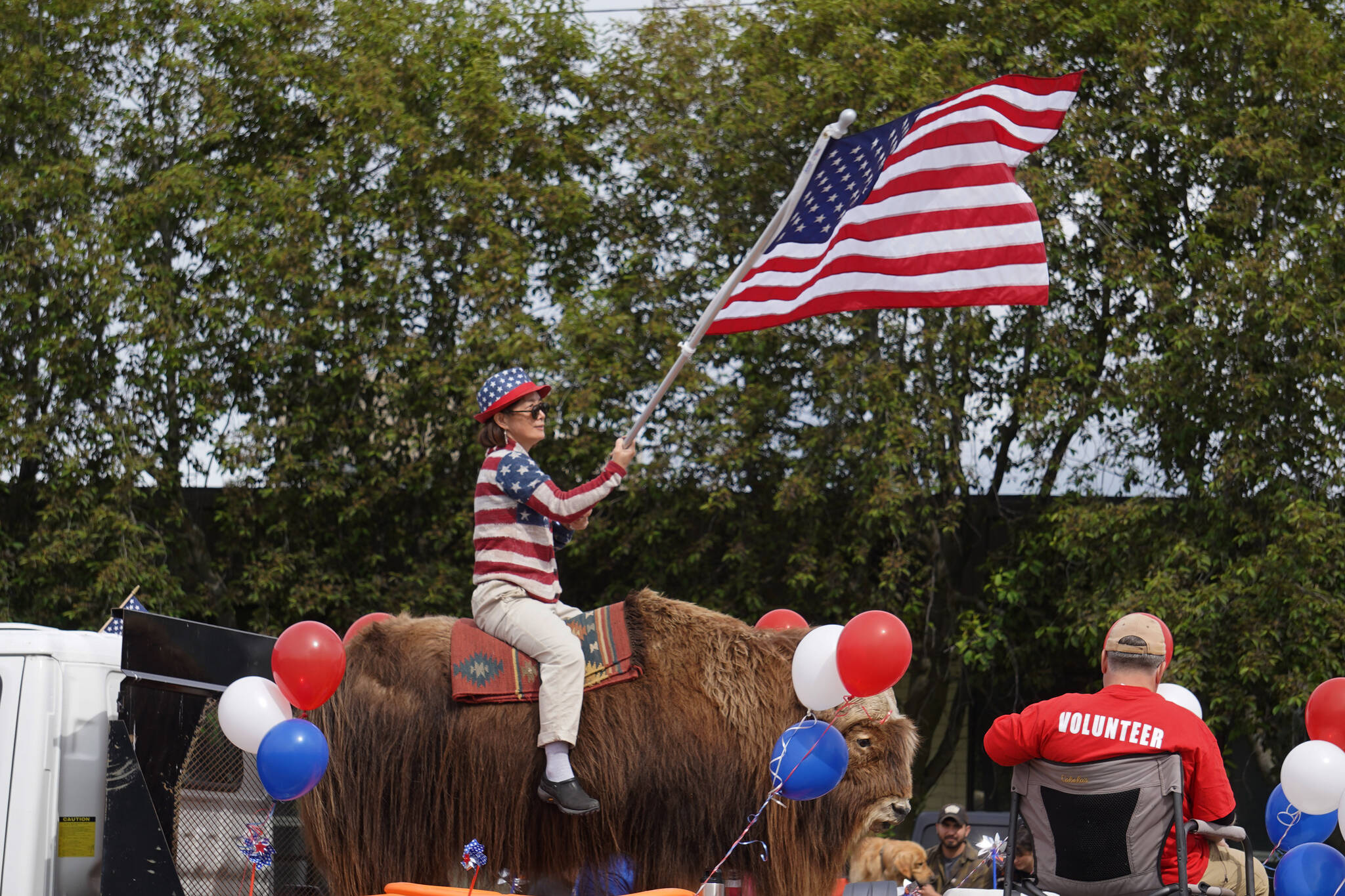 A woman waves a flag from atop a large animal as part of the Fourth of July Parade down the Kenai Spur Highway in Kenai, Alaska on Tuesday, July 4, 2023. (Jake Dye/Peninsula Clarion)