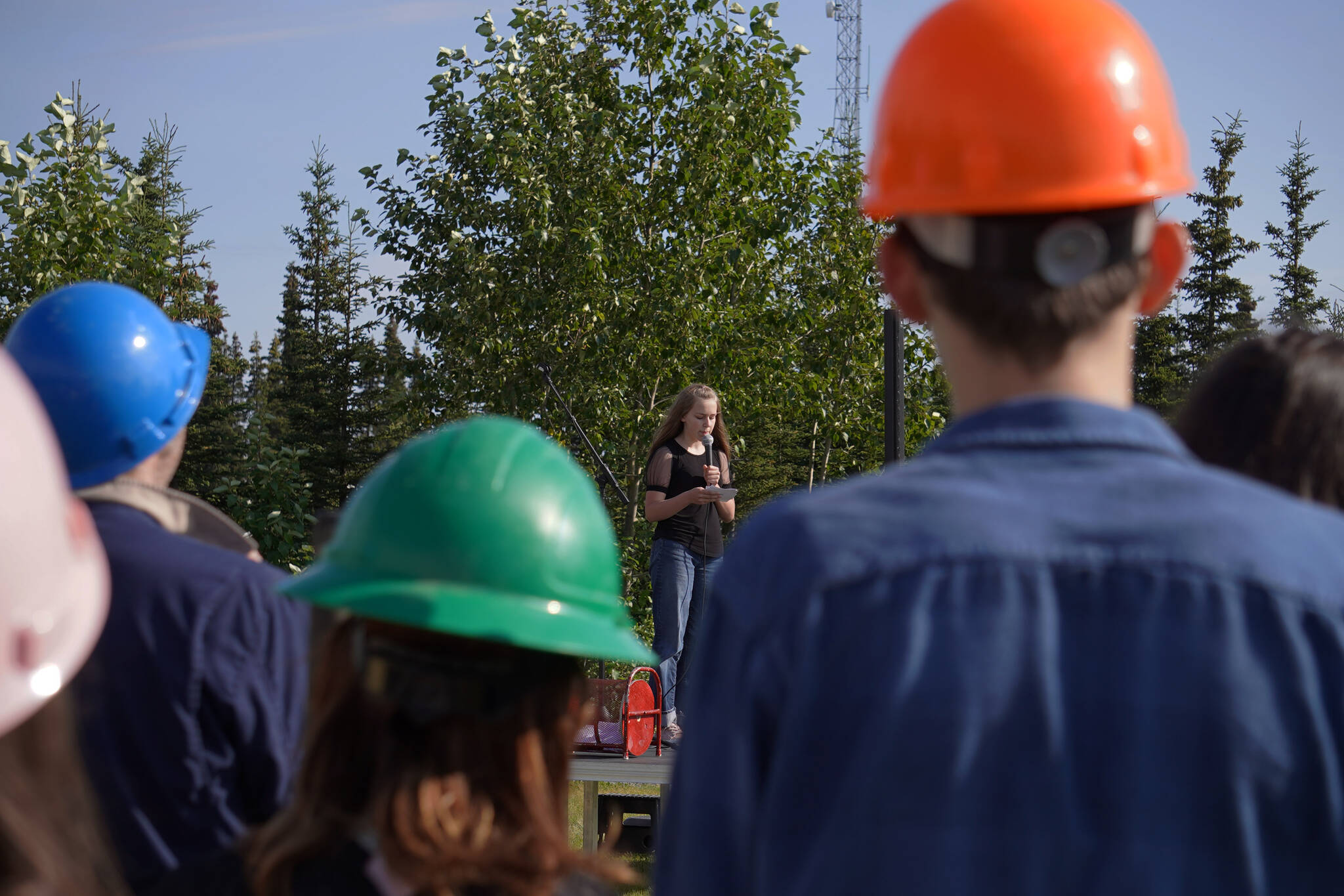Rows of Triumvirate Theatre performers, equipped with hard hats, listen to a speech by fellow actor Cora Frazier during a ceremonial groundbreaking for the future home of Triumvirate Theatre at Daubenspeck Family Park in Kenai, Alaska, on Saturday, Aug. 19, 2023. (Jake Dye/Peninsula Clarion)