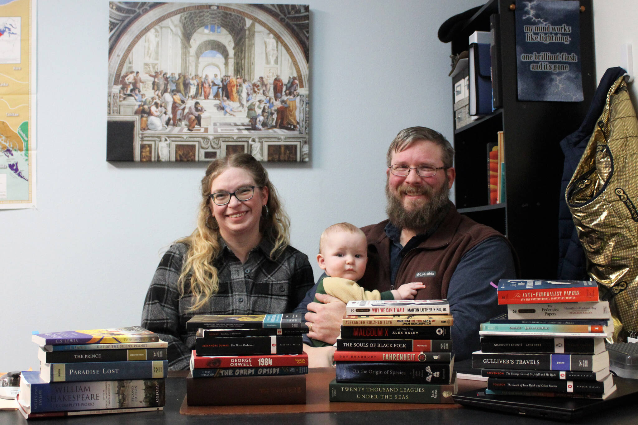 From left: Kylie Wilcox, Bernard Wilcox and Milton Wilcox sit behind the books Milton teaches at Kenai Classical School on Wednesday, Dec. 27, 2023 in Kenai, Alaska. Kylie’s New Year’s resolution for 2024 is to read all of the book’s on Milton’s high school syllabus. (Ashlyn O’Hara/Peninsula Clarion)