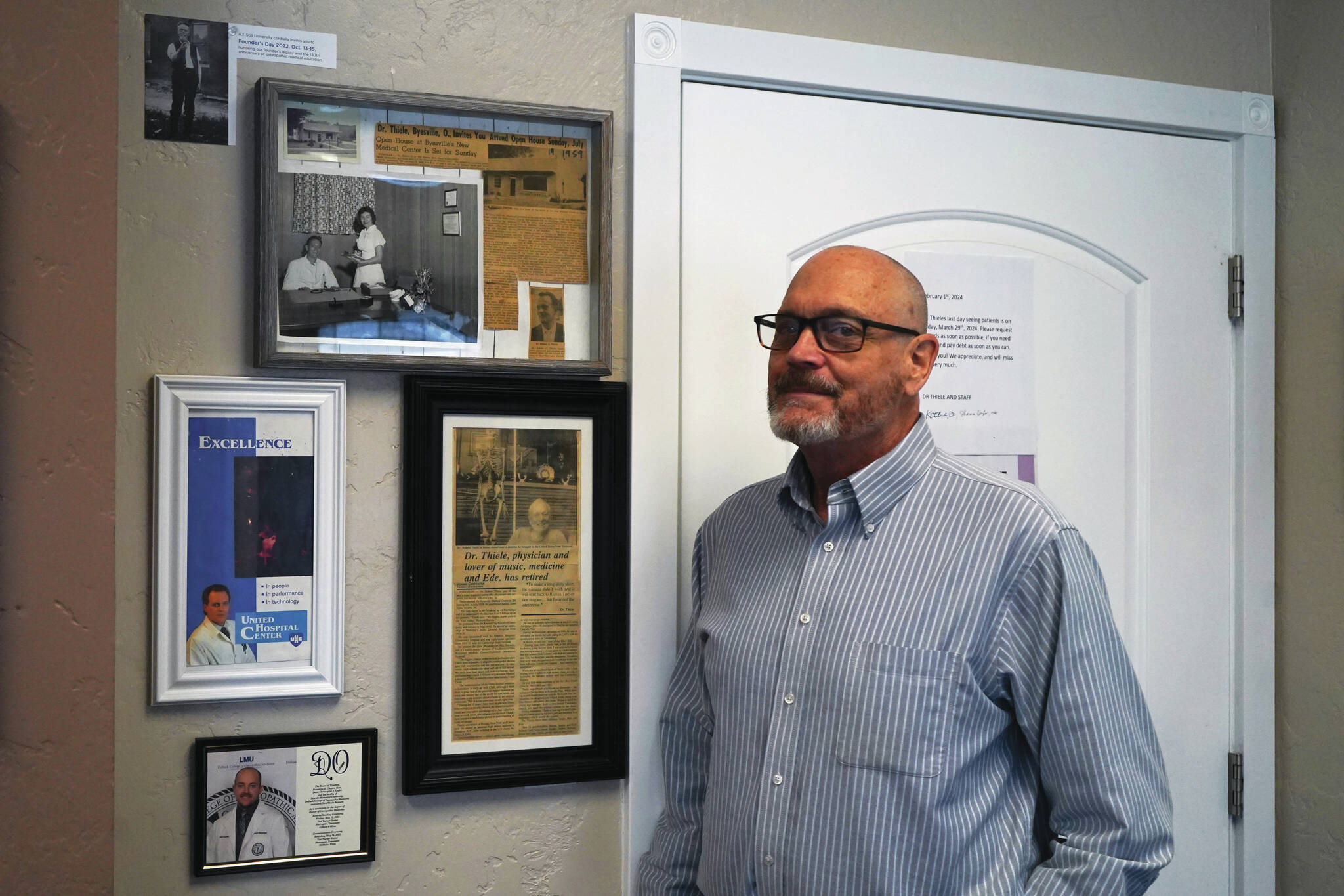 Dr. Kim Thiele stands by a wall of newspaper clippings and images of family members and precursors in his office near Kenai on Monday. (Jake Dye/Peninsula Clarion)