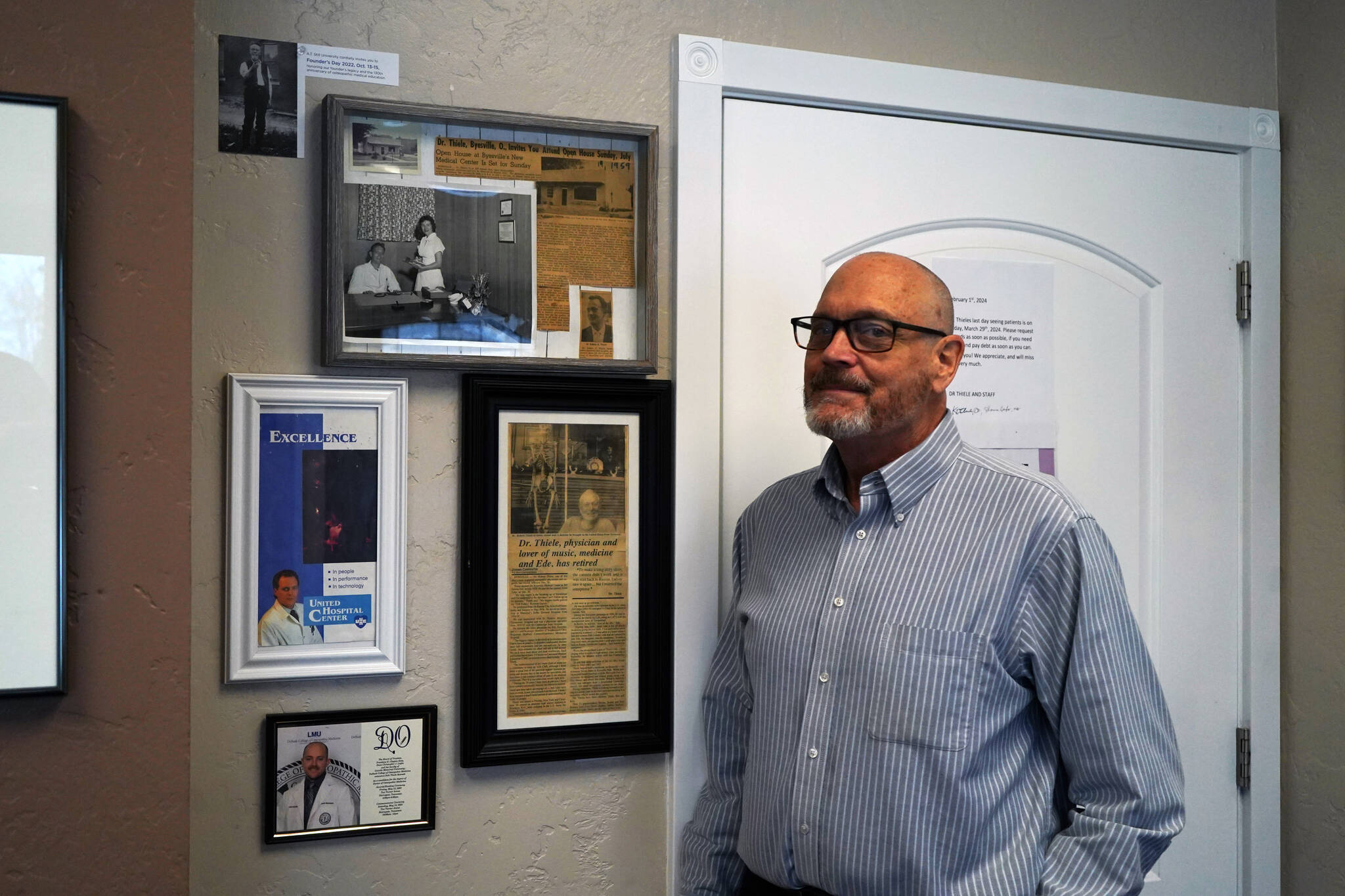 Dr. Kim Thiele stands by a wall of newspaper clippings and images of family members and precursors in his office near Kenai, Alaska, on Monday, Feb. 21, 2024. (Jake Dye/Peninsula Clarion)
