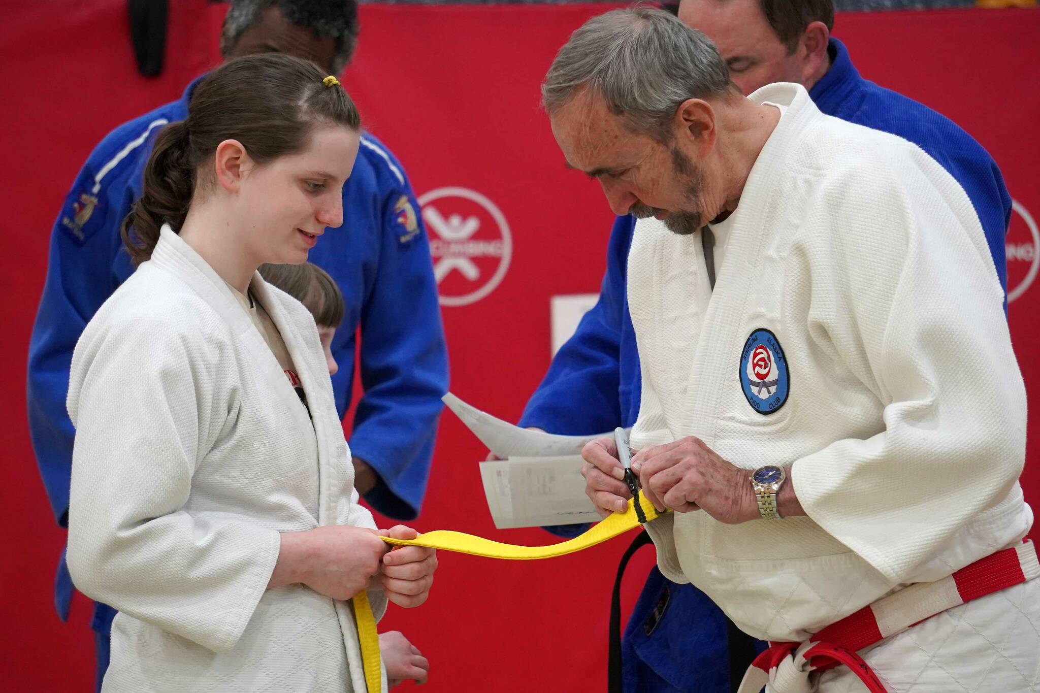 Arianna Jicha watches as Sensei Bob Brink adds a bar to her yellow belt at the Sterling Judo Club’s end of season awards night at Sterling Elementary School in Sterling, Alaska, on Thursday, May 15, 2025. (Jake Dye/Peninsula Clarion)