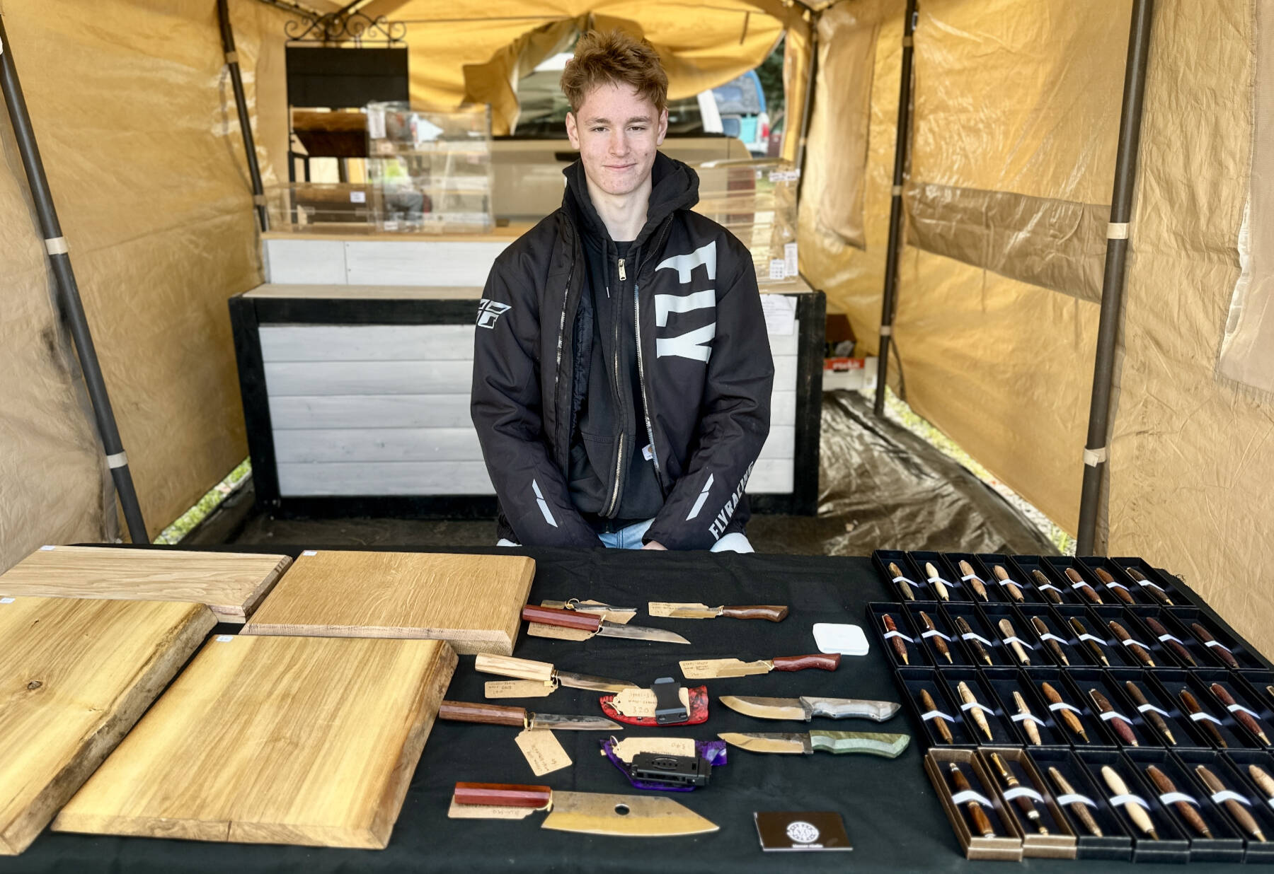 Jaxon Bourne sits in his booth at the Homer Farmers Market with his handmade knives, pens and cutting boards on Saturday, Sept. 13, 2025, in Homer, Alaska. Photo by Christina Whiting