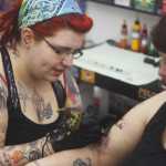 Photo by Kelly Sullivan/ Peninsula Clarion Sam Hayes works on a black and white image of a cat sitting beside a tombstone just below Lori Salyers shoulder, Saturday, March 14, 2015, at Ink Works in Kenai, Alaska.