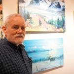 Bill Smith stands by a John Fenske painting, top, that his children bought for him. Fenskes retrospective show opened on First Friday, April 5, 2019, at Kachemak Bay Campus in Homer, Alaska. (Photo by Michael Armstrong/Homer News)