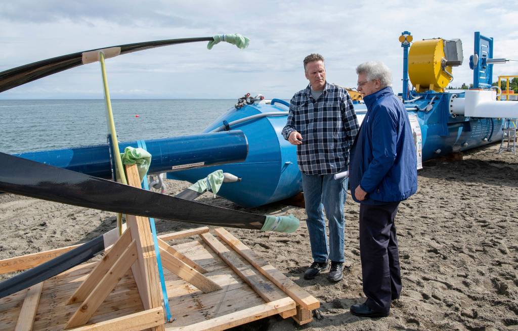 In this Tuesday, July 16, 2019, photo released by the Alaska Governors office, Gov. Mike Dunleavy, left, talks with ORPC CEO, Co-Founder, and Chairman Chris Sauer in front of a Riv-Gen Power System turbine on the bank of the Kvichak River in Igiugig, Alaska. A tiny Alaska Native village is adopting an emerging technology to transform the power of a local river into a renewable energy source. (Austin McDaniel/Alaska Governors Office via AP)