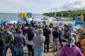 In this Tuesday, July 16, 2019, photo released by the Alaska Governors office, Gov. Mike Dunleavy, center rear, speaks in front of a Riv-Gen Power System turbine on the bank of the Kvichak River in Igiugig, Alaska. A tiny Alaska Native village is adopting an emerging technology to transform the power of a local river into a renewable energy source. (Austin McDaniel/Alaska Governors Office via AP)